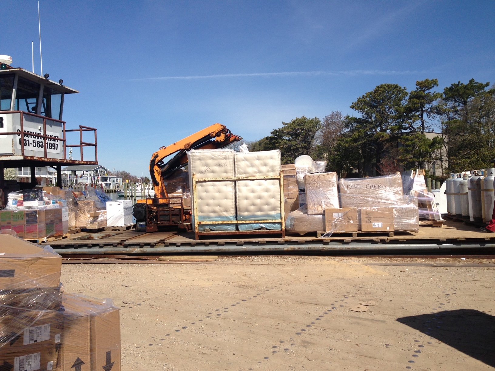 Storage yard with pallets of furniture and boxes, a crane, and trees in the background.