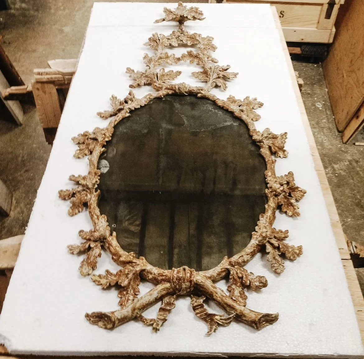 An ornate oval mirror with a detailed wooden frame resembling tree branches, laying on a white surface in a workshop.