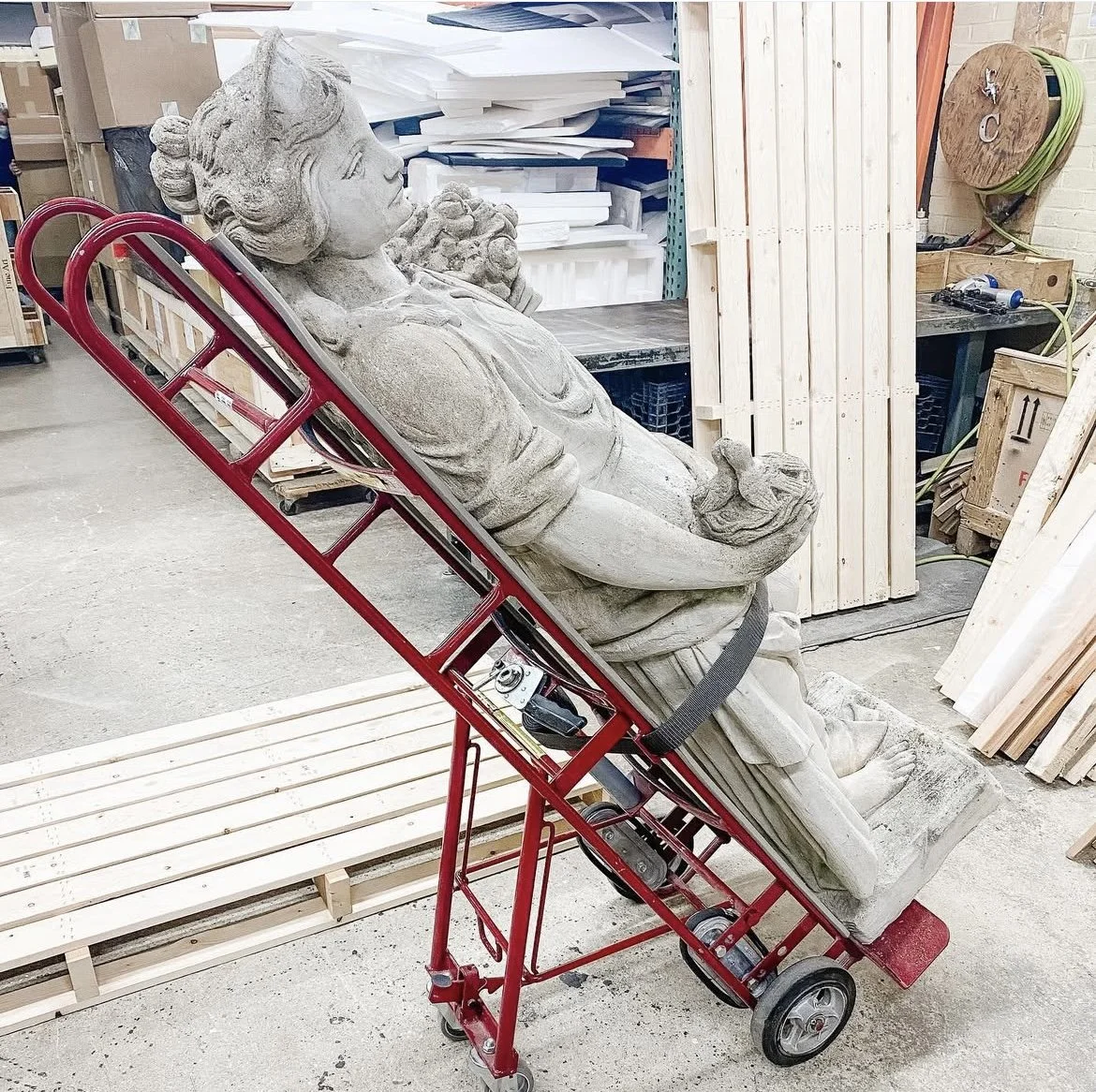 Stone statue of a woman with wings, lying on a red dolly in a woodworking workshop.