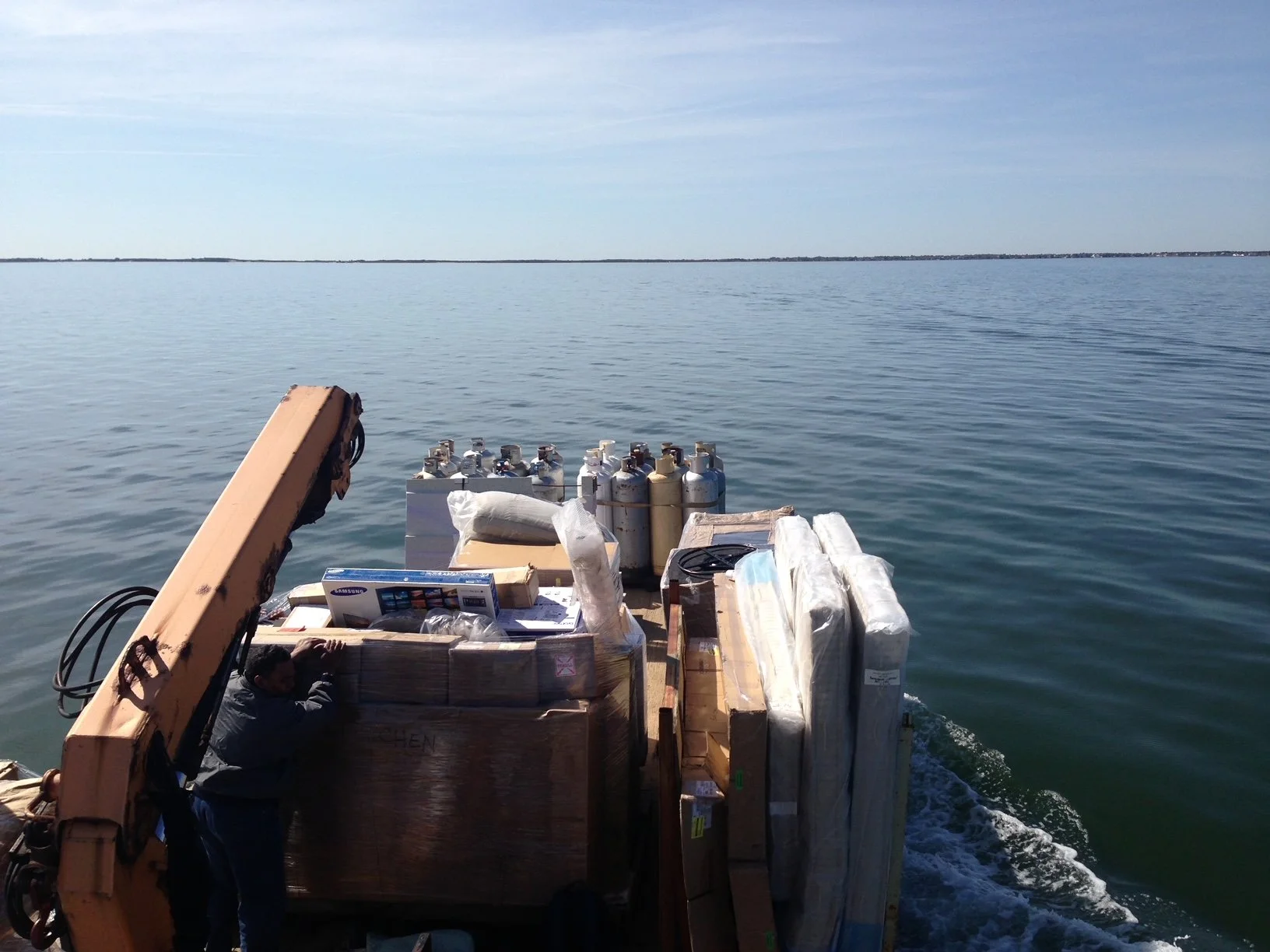 Cargo on a boat, including boxes, gas tanks, wrapped items, and an individual, on a body of water with a distant shoreline visible under a clear sky.