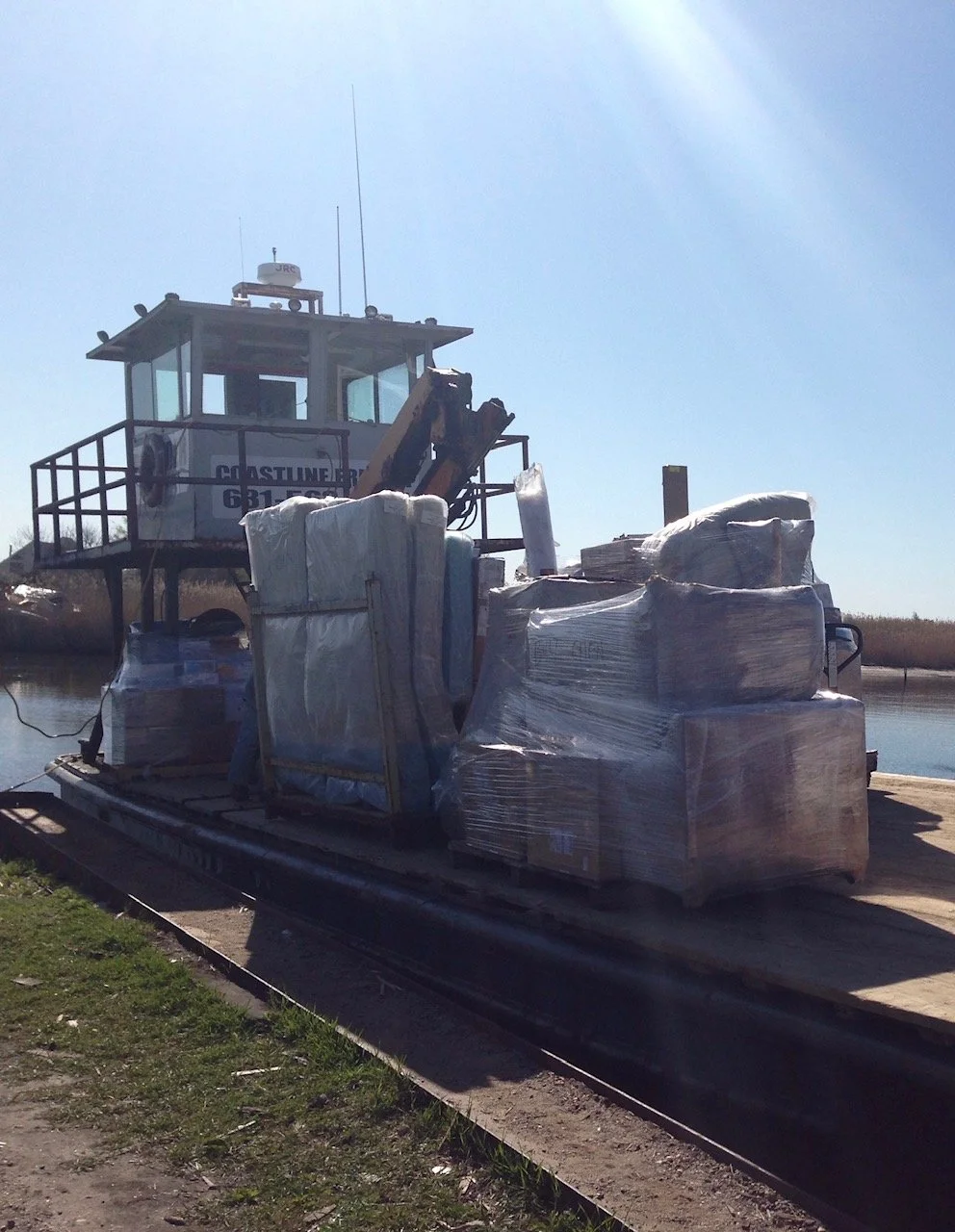A barge loaded with wrapped pallets and luggage, with a watercraft's control cabin in the background.