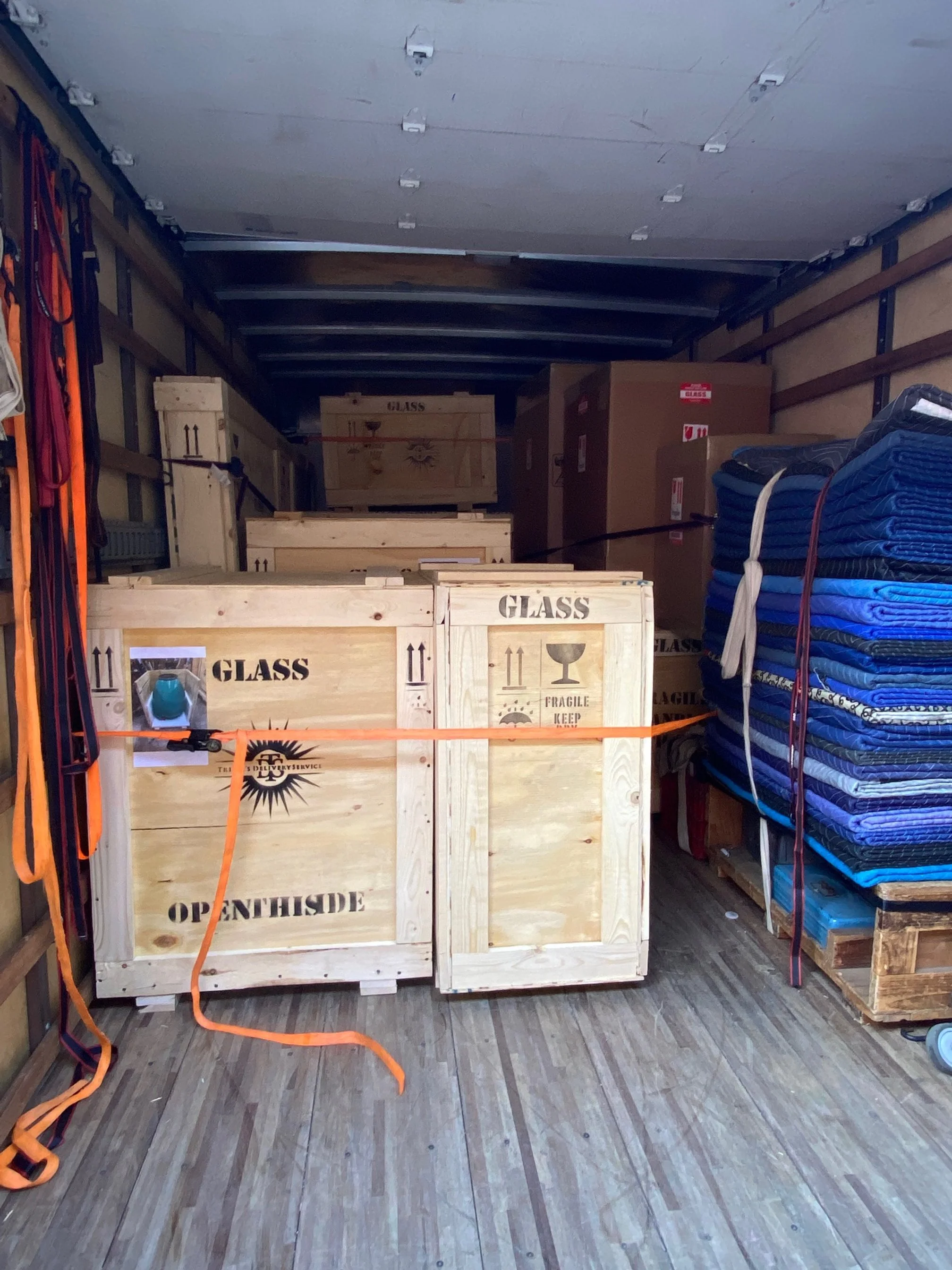 Wooden crates labeled 'GLASS' and 'OPENHIDE' inside a truck, with stacked blue tarps on the right and orange straps securing the cargo.