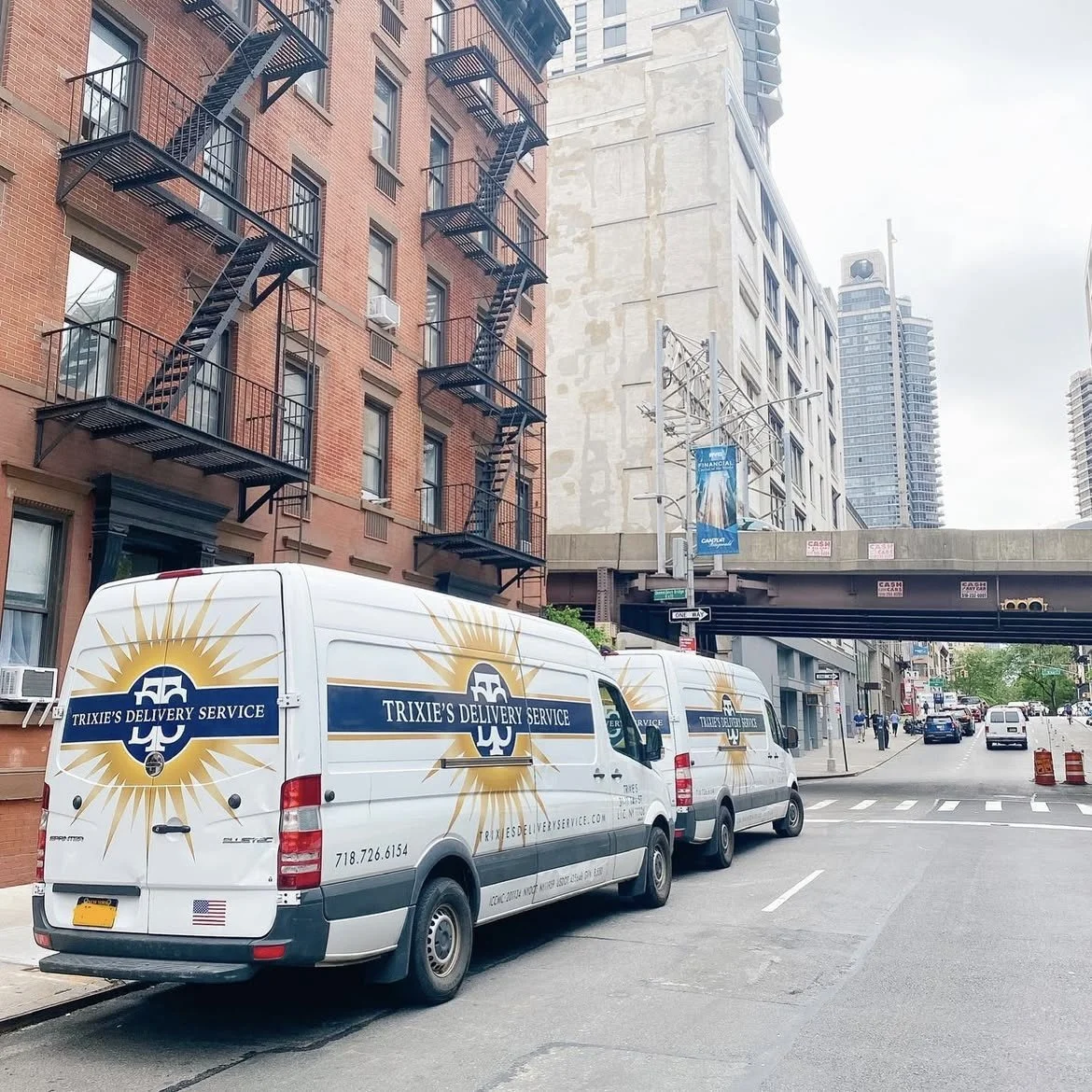 Two white delivery vans with a logo and text that reads 'Trixie's Delivery Service' are parked on the side of a city street. There are brick and concrete buildings, a fire escape, and a bridge overhead.