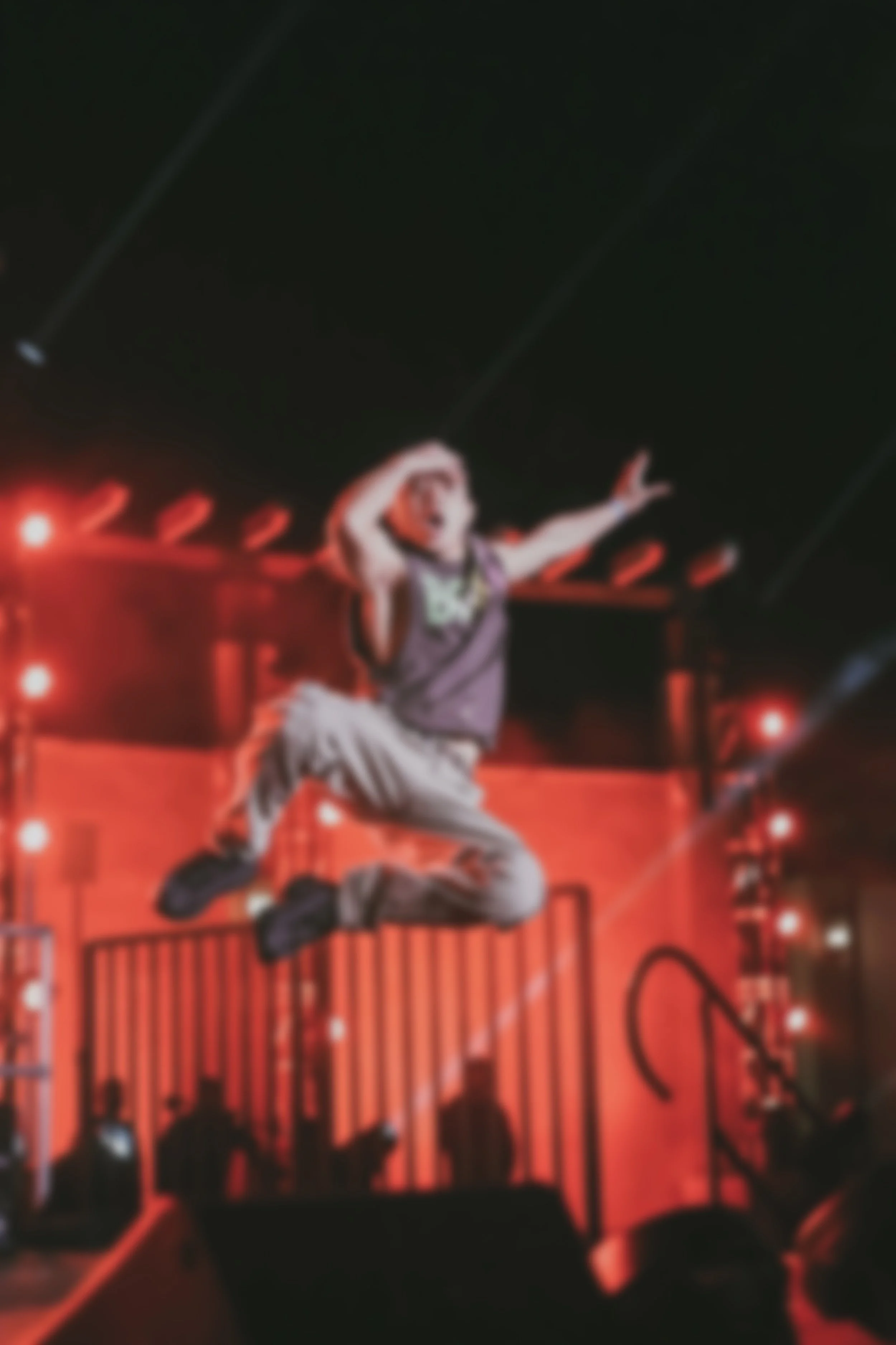 A person jumping in the air on a stage with red lighting, with their hand on their head and wearing casual clothes, during a performance or concert.