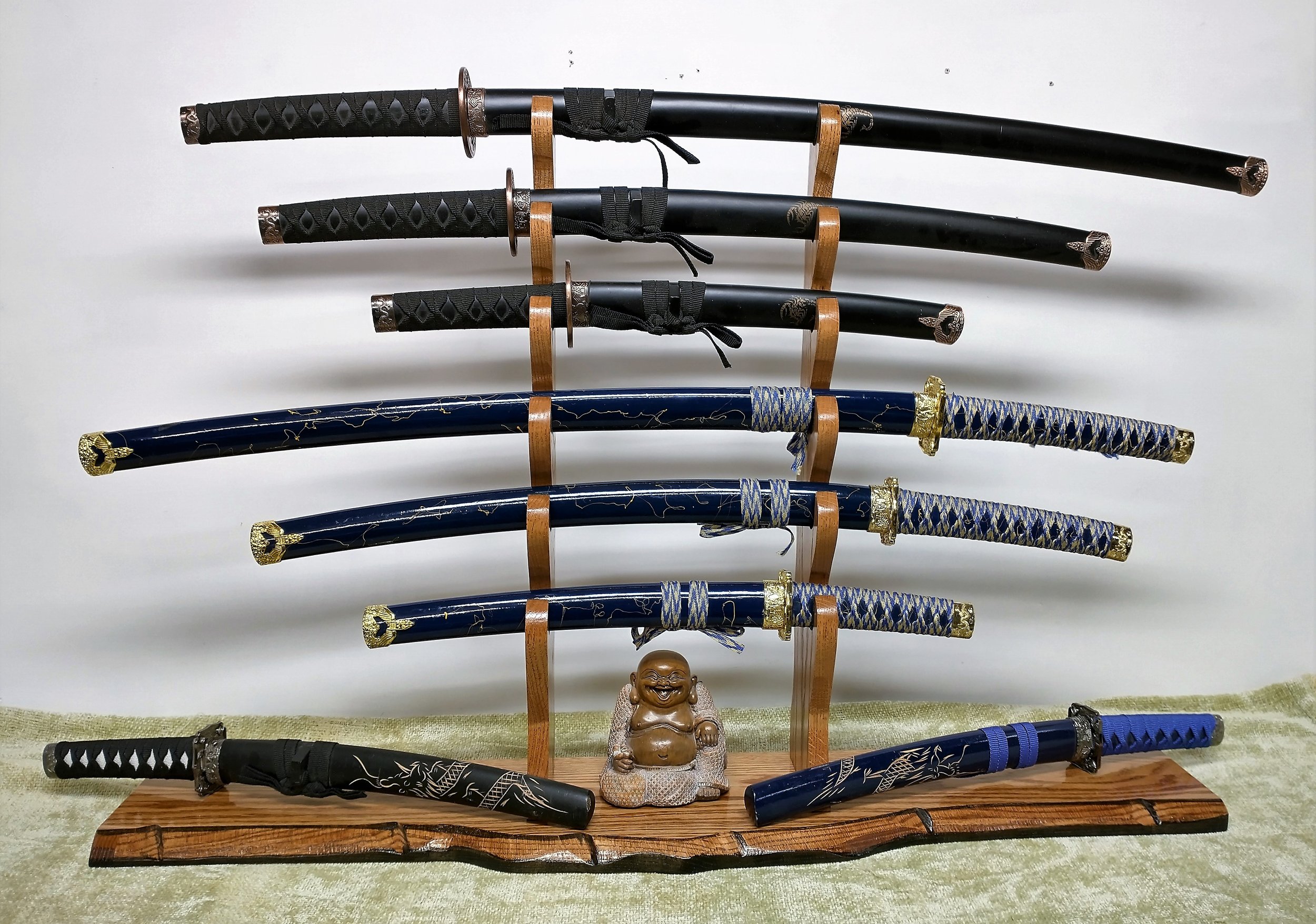 Display of eight Japanese swords, five with black blades and three with blue blades featuring gold accents, arranged on wooden stands against a plain background. A small Buddha statue is placed at the bottom center.