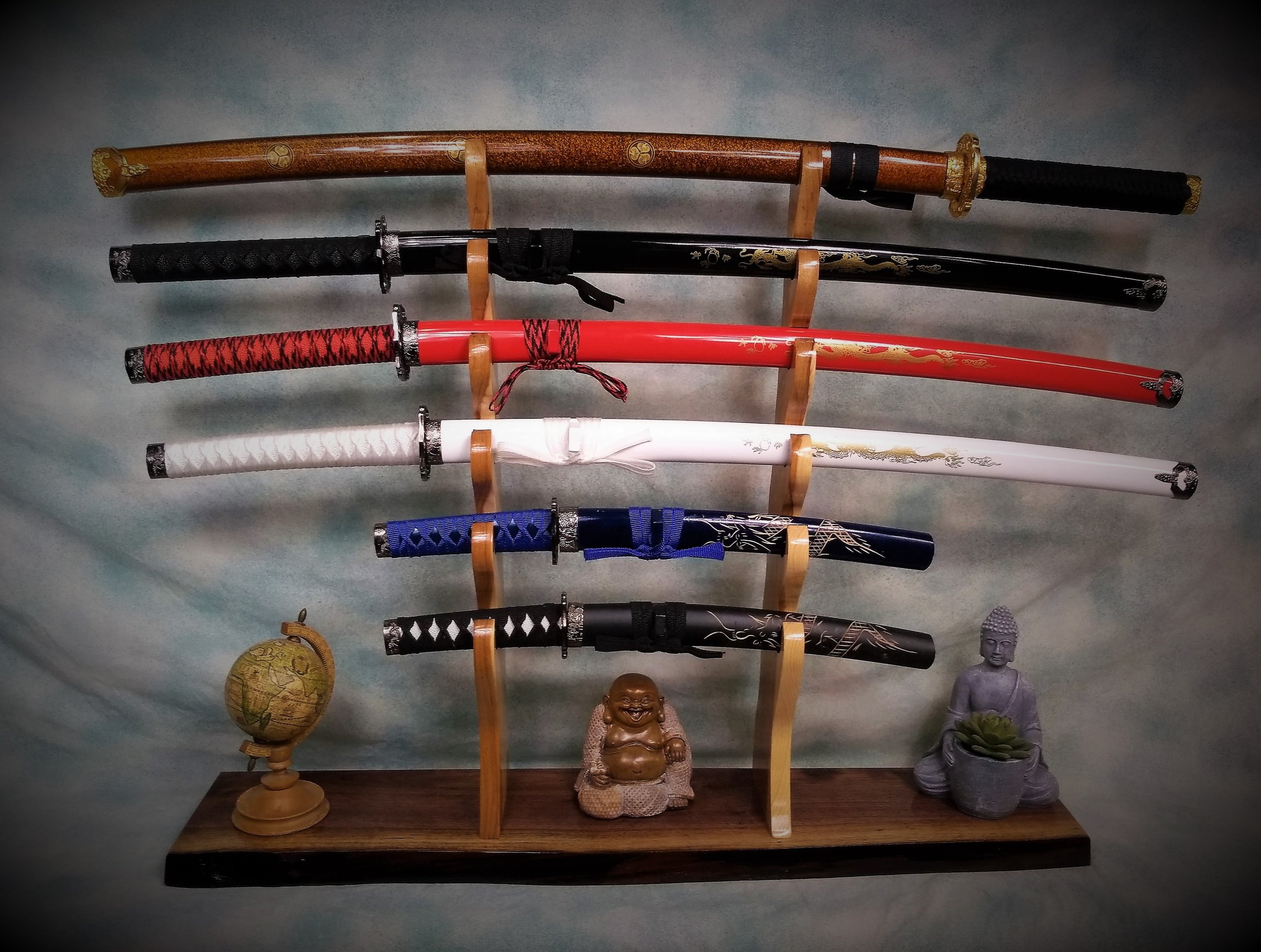 A display of seven decorative katana swords on a wooden stand, with a small globe, a laughing Buddha figurine, and a seated Buddha statue with a lotus flower on a wooden surface.