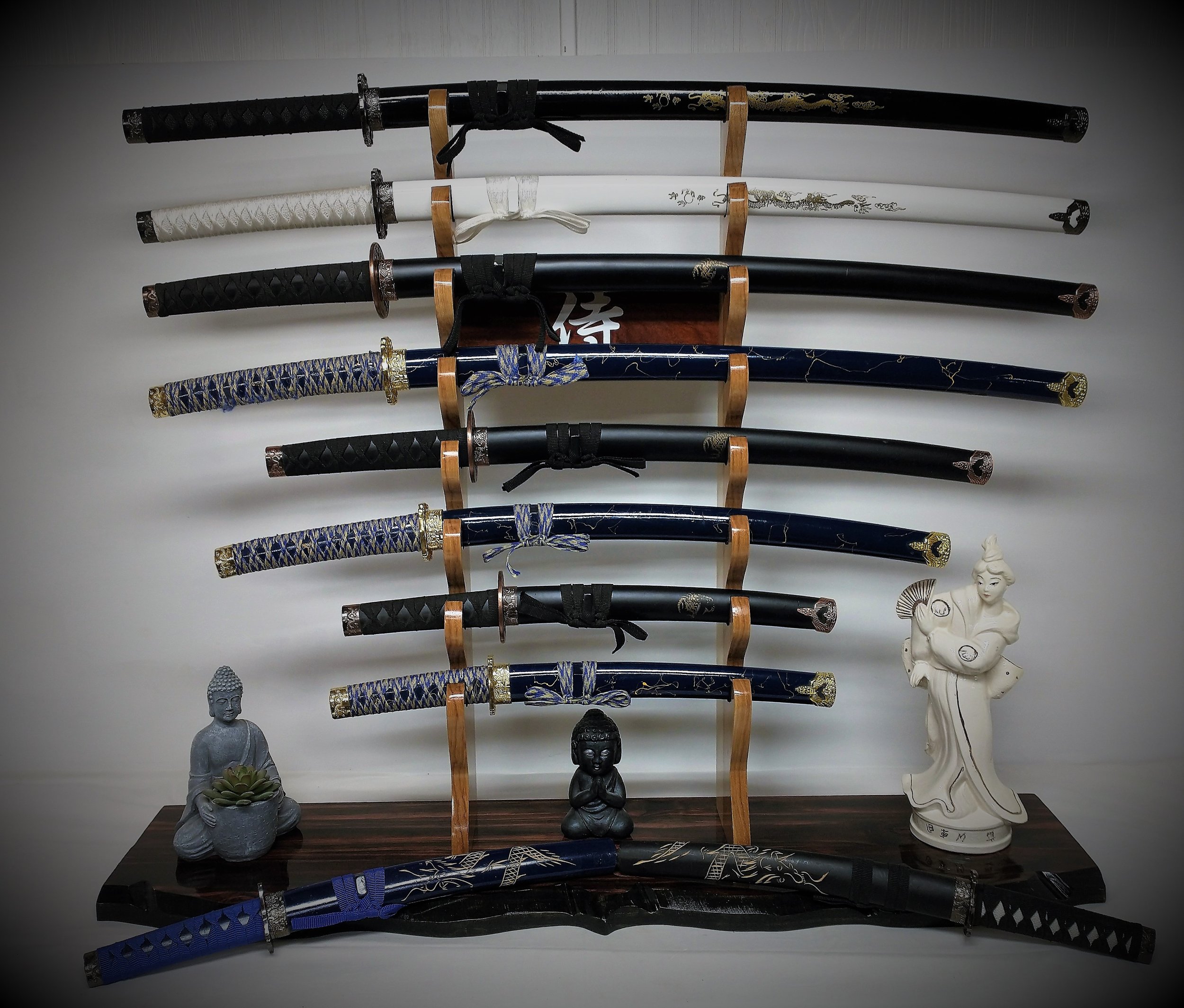 A display of nine Japanese swords mounted on a wall with decorative scabbards and handles, accompanied by Buddhist statues and a wooden stand.