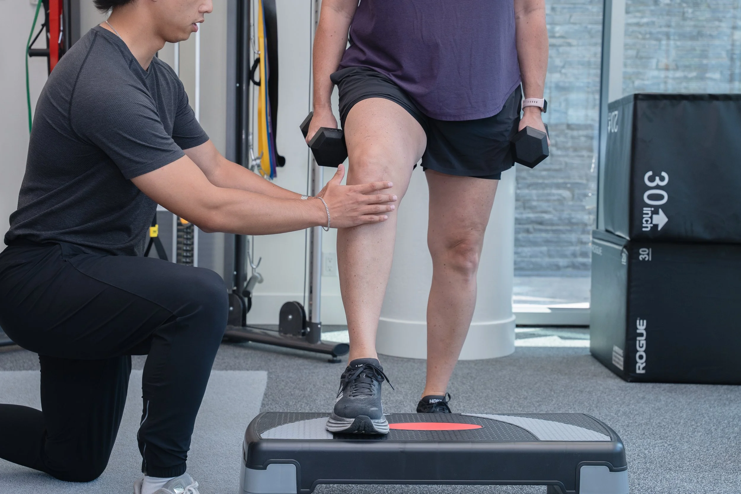 A person performing step exercises with weights while a trainer assists and provides support during the workout.