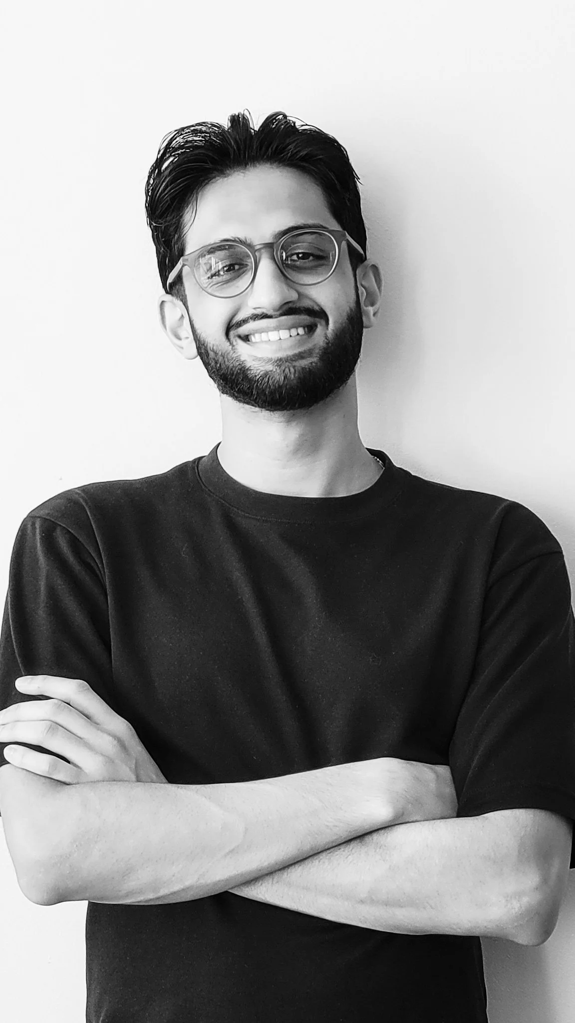Black and white portrait of a young man with glasses, a beard, and messy hair, smiling with arms crossed, standing against a plain background.