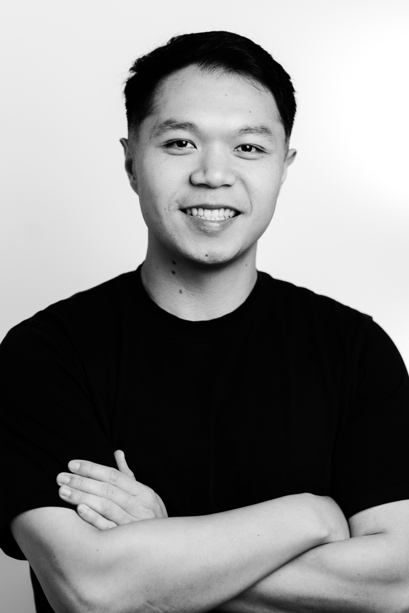 Black and white portrait of a young man smiling, with short dark hair, crossed arms, wearing a black t-shirt, against a plain light background.