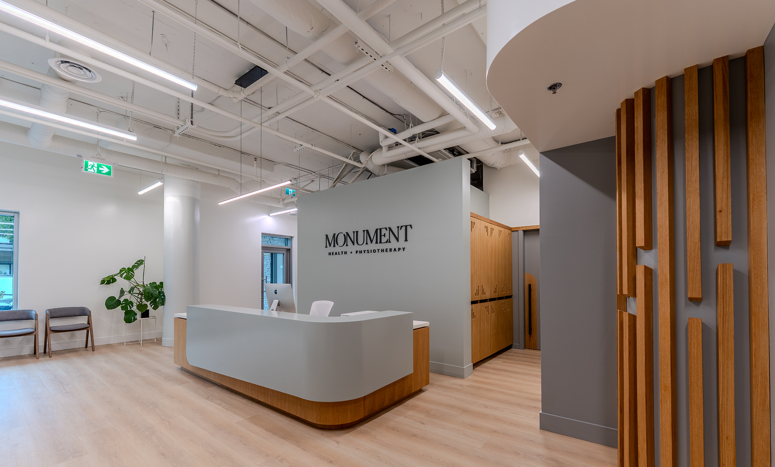 Modern clinic reception area with white walls, wooden accents, a curved reception desk, a small seating area with chairs, a large plant, and signage that reads 'Monument Health + Physiotherapy'.