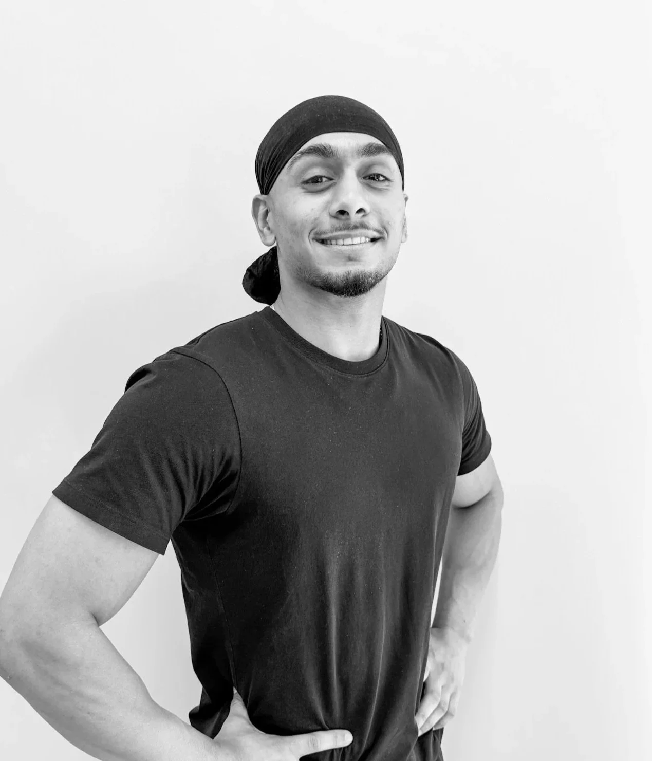 A man with a dark head covering, black t-shirt, smiling and posing with hands on hips, standing against a plain white wall.