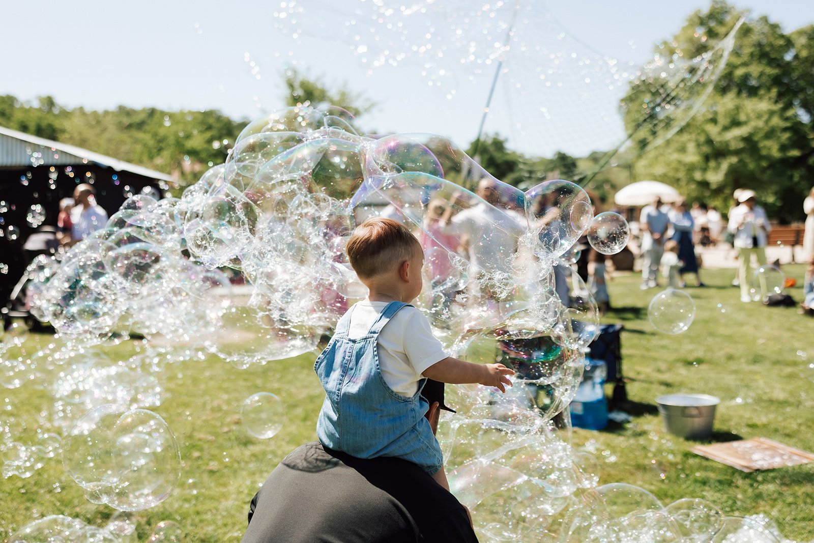 Young child wearing denim overalls playing with soap bubbles at an outdoor event on a sunny day, with people in the background enjoying the gathering.