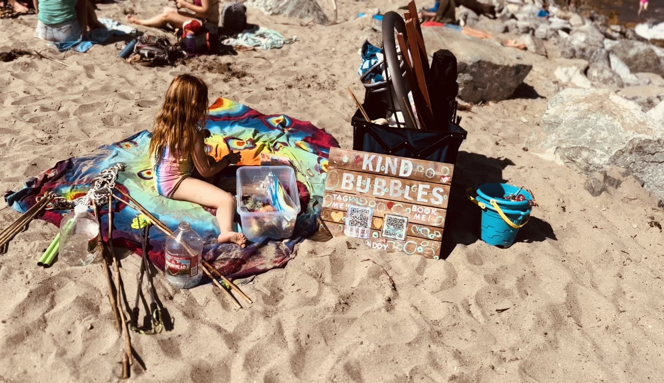 A young girl sitting on a colorful blanket on the sandy beach, playing with bubbles. Next to her is a plastic container and a signboard that reads 'KIND BUBBLES' with QR codes and a book icon. There are buckets, toys, rocks, and beach items scattered