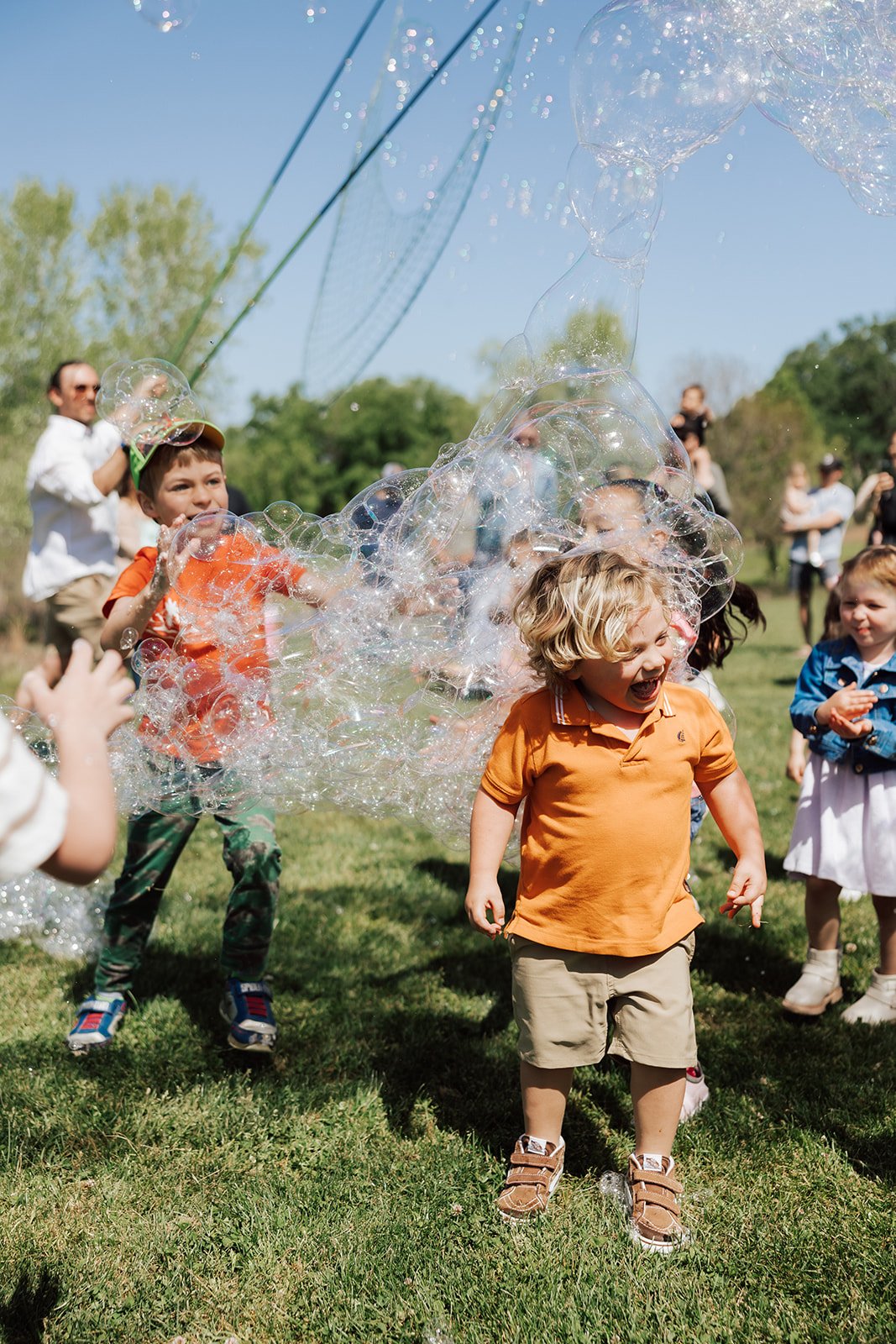 Children playing with bubbles outdoors on a sunny day, with a green park and blue sky in the background.