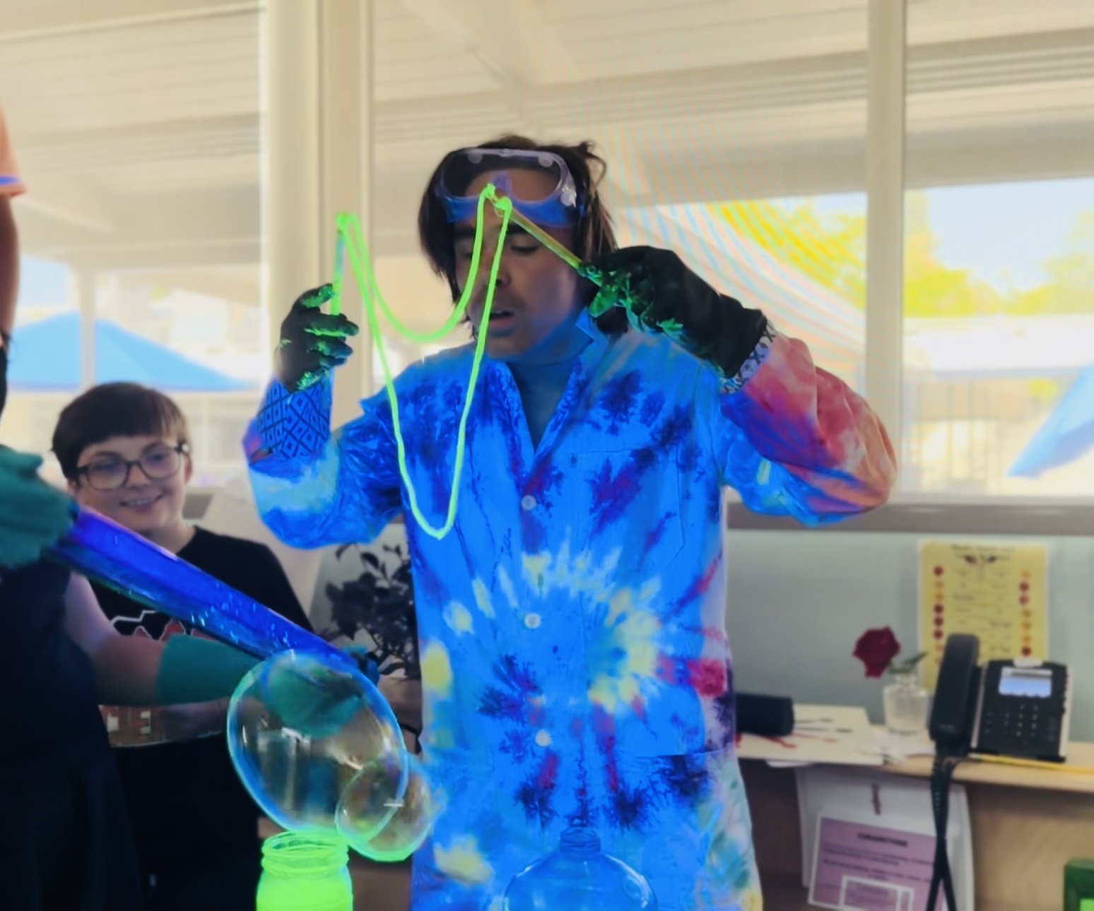 Person creating glowing bubbles using a bubble mixture and a glow stick, with a child watching and smiling in the background.