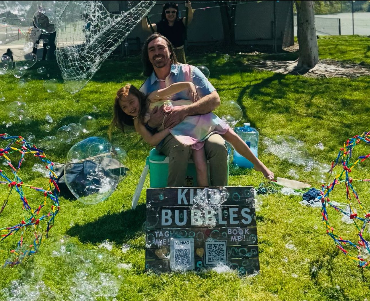 A man and a young girl are sitting on a green lawn, surrounded by bubbles. The man is holding the girl, who is smiling and leaning into him. There are two colorful bubble frames on either side of the scene, and a sign that reads "KID BUBBLES" with QR