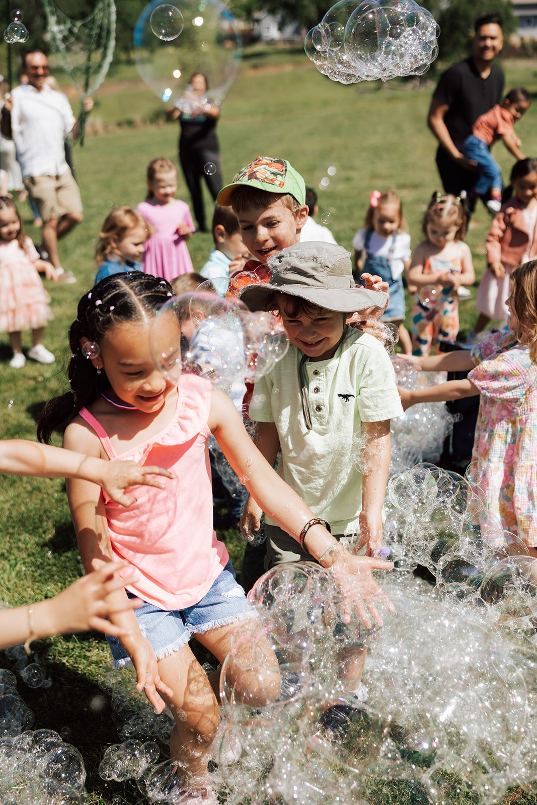 Children playing with soap bubbles outdoors at a sunny daytime birthday party or gathering, surrounded by adults and other kids.