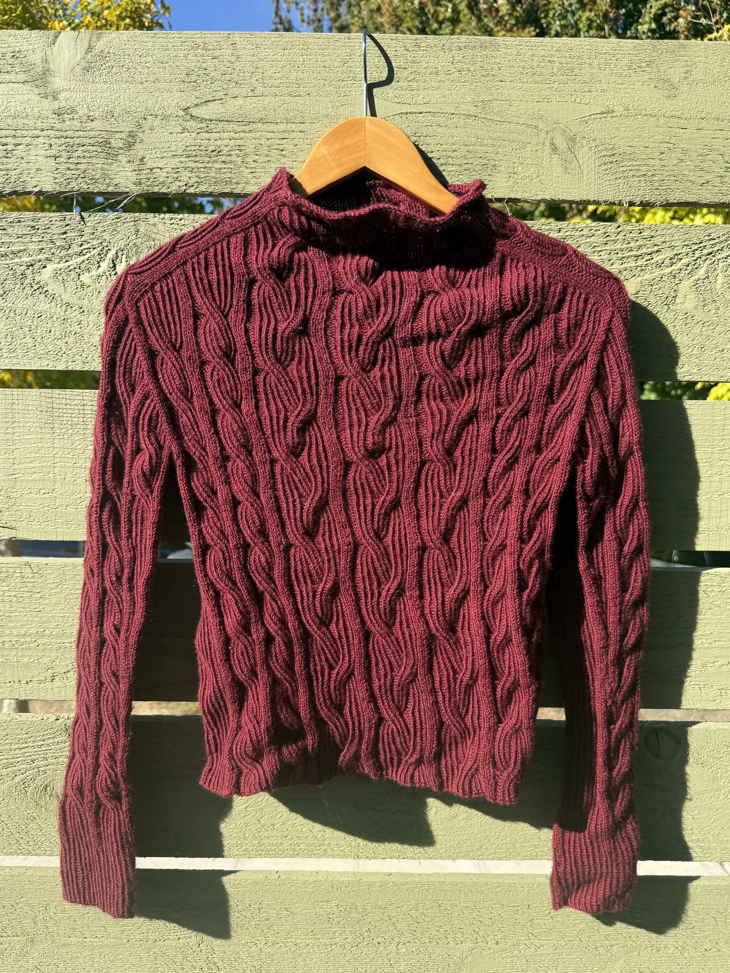 A maroon, cable-knit turtleneck sweater hanging on a wooden hanger outside against a light green wooden fence.