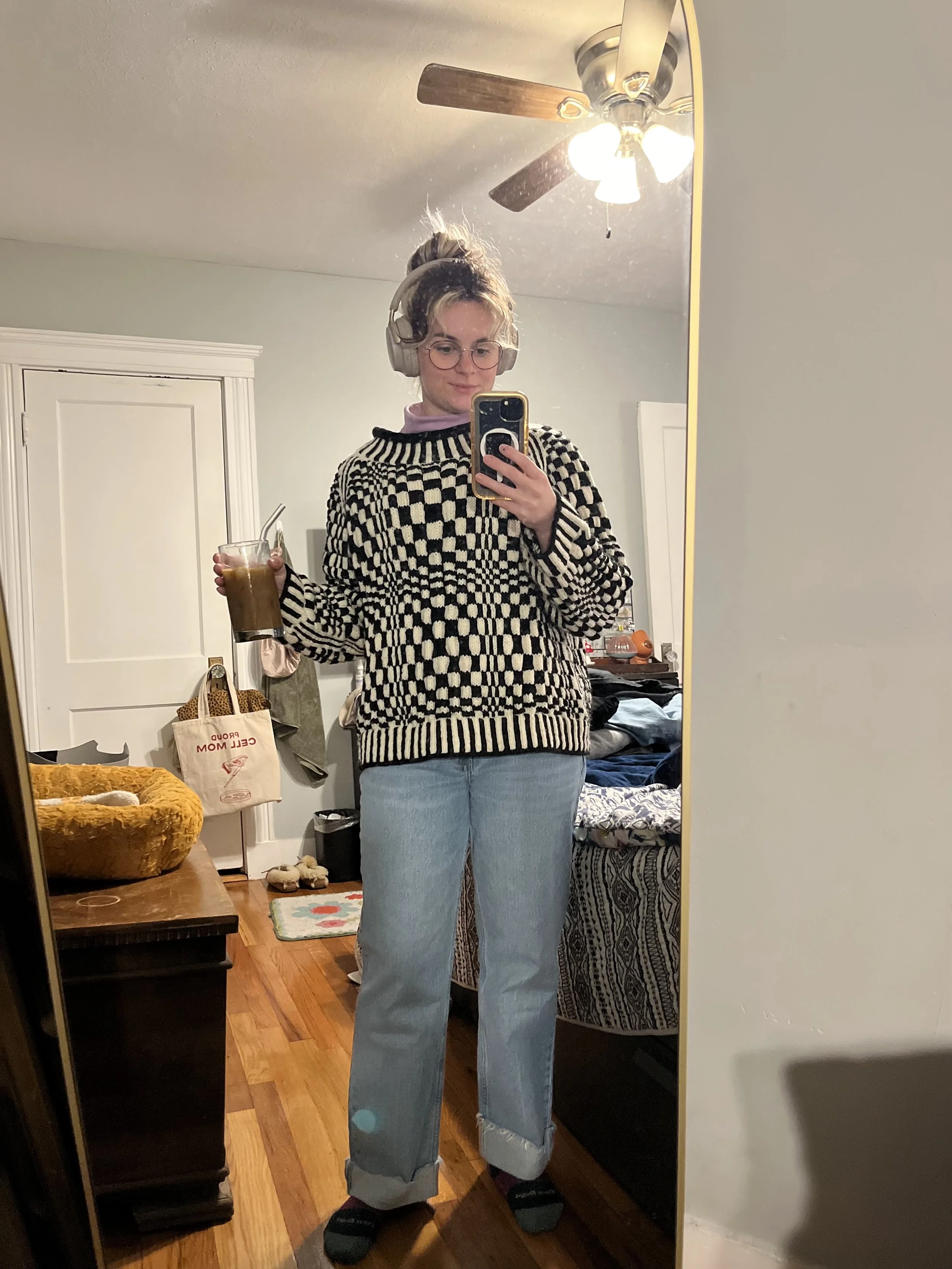A young woman with glasses, wearing headphones, a patterned black and white sweater resembling an optical illusion, light blue jeans, and slippers, taking a mirror selfie in a bedroom while holding a cup of iced coffee.