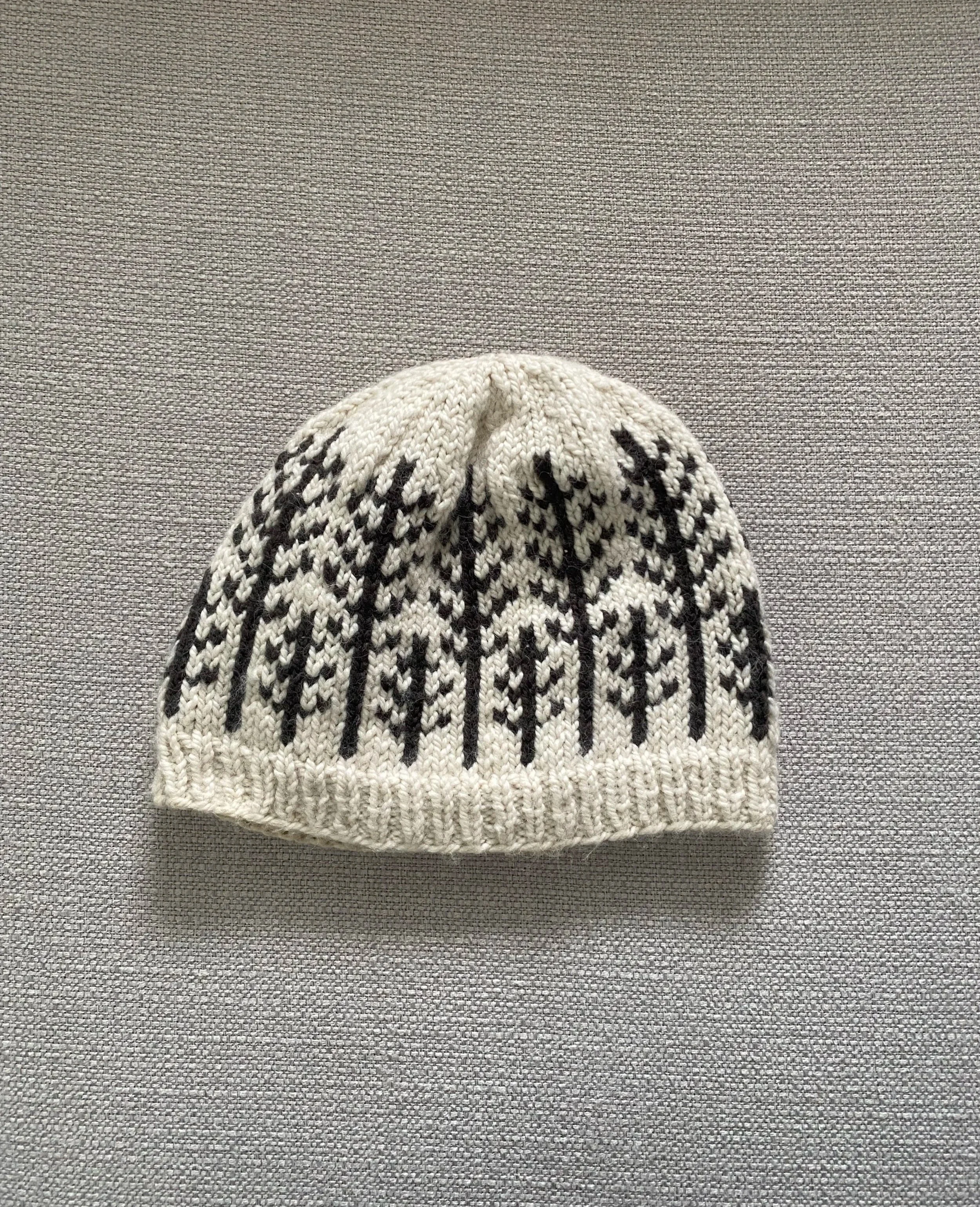 A knitted white beanie hat with black tree patterns, placed on a gray textured surface.