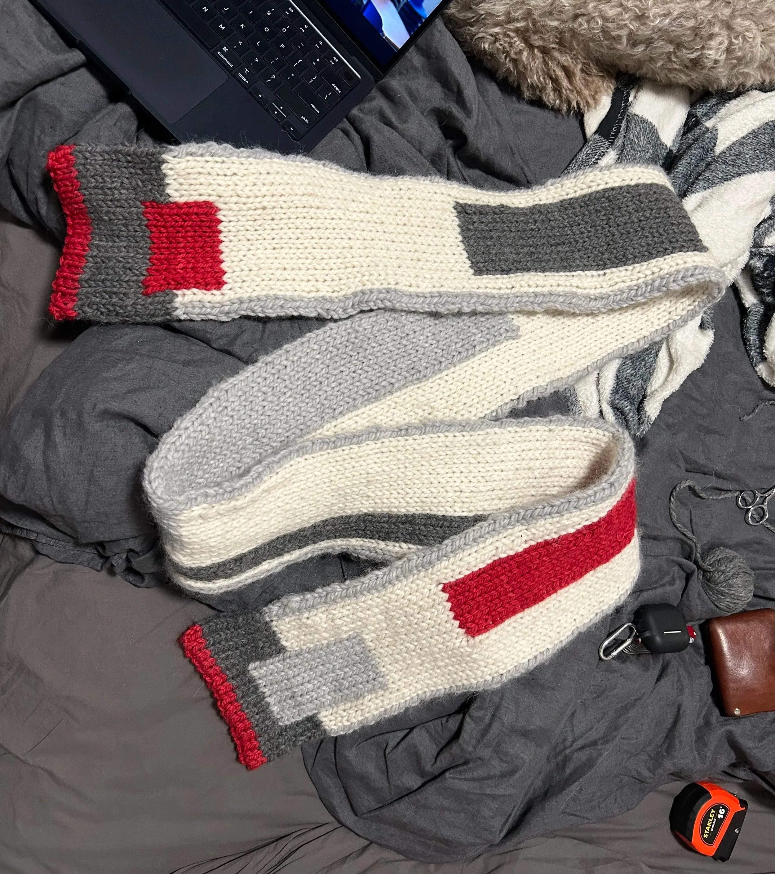 Knitted scarf with geometric color blocks in red, gray, white, and cream, laid out on a dark gray bed spread next to a laptop and a pair of scissors.