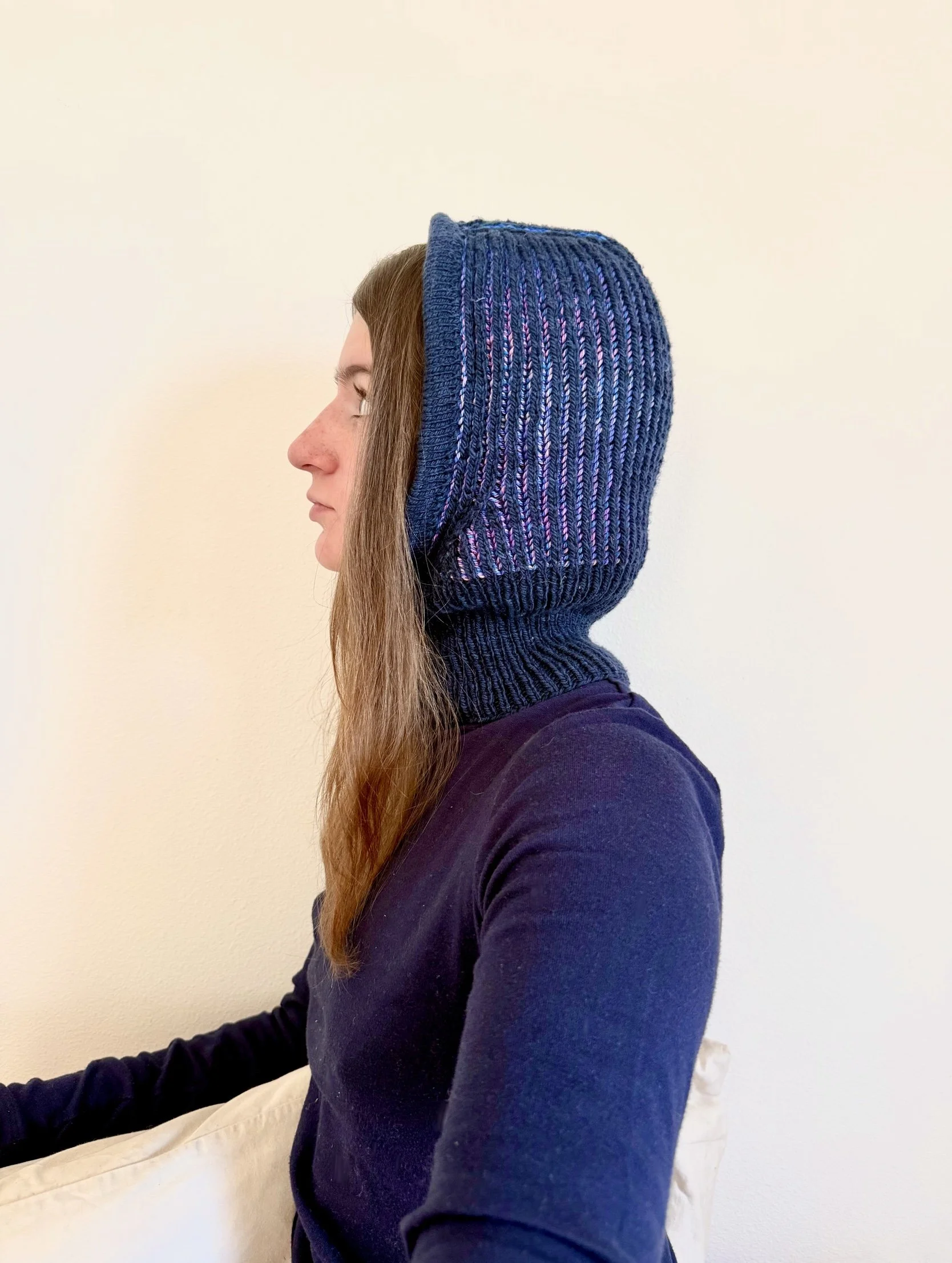 Woman in profile wearing a blue knit hood with striped pattern and a navy blue long sleeve shirt, sitting against a plain white wall.