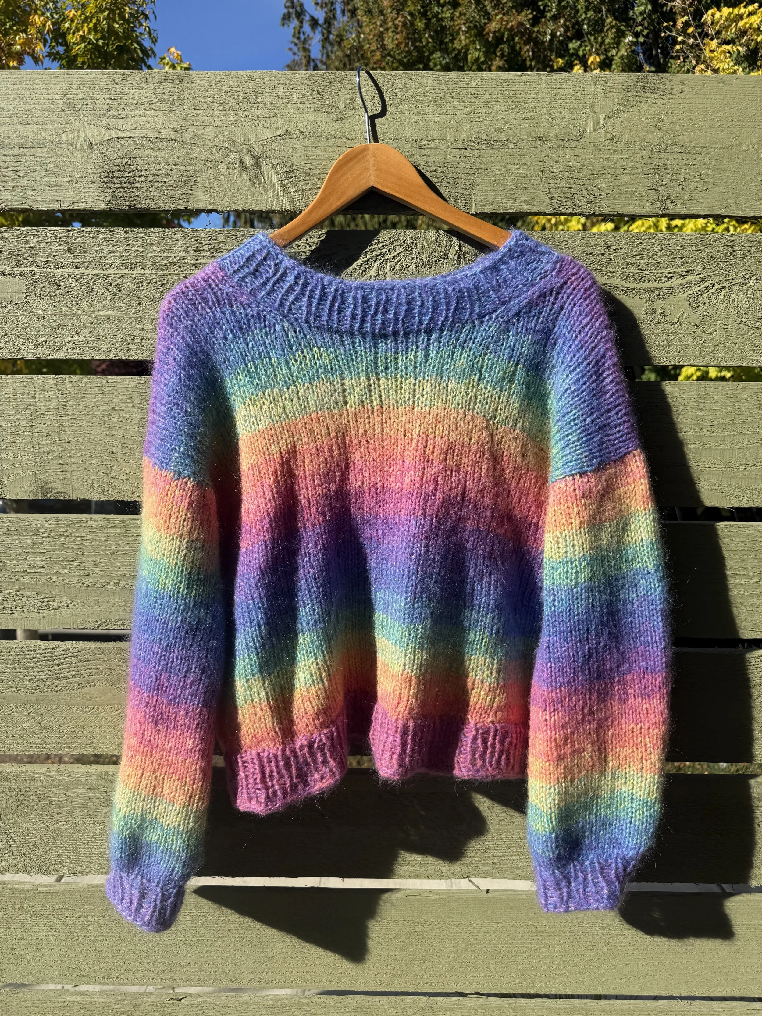 A rainbow-colored knitted sweater hanging on a wooden hanger against a green wooden fence with a clear blue sky above.