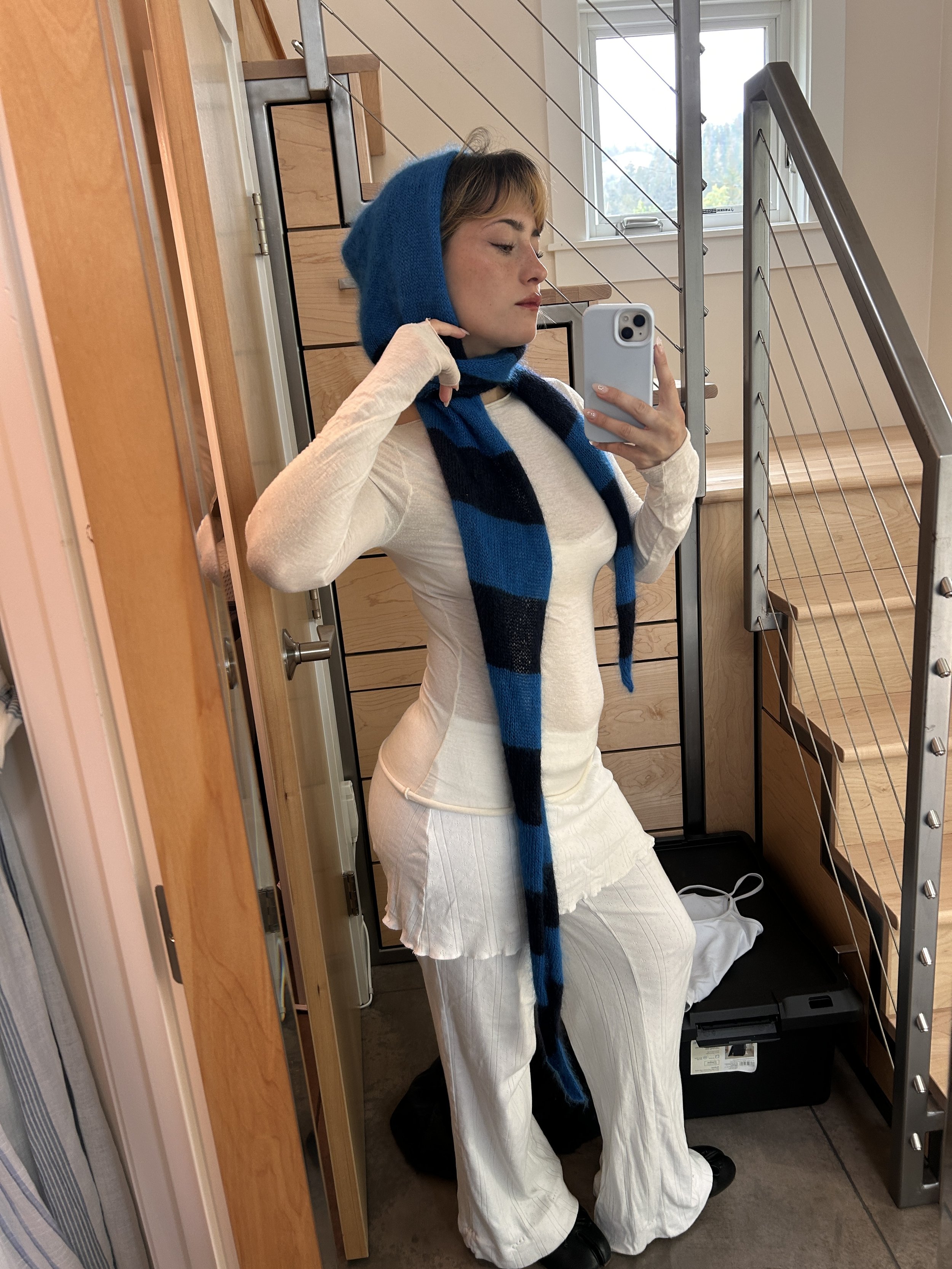 Young woman taking a mirror selfie indoors, wearing a blue and black striped scarf and a hood, a light-colored long-sleeve top, matching light-colored pants, and black shoes. She stands near wooden furniture and stairs with metal railings.