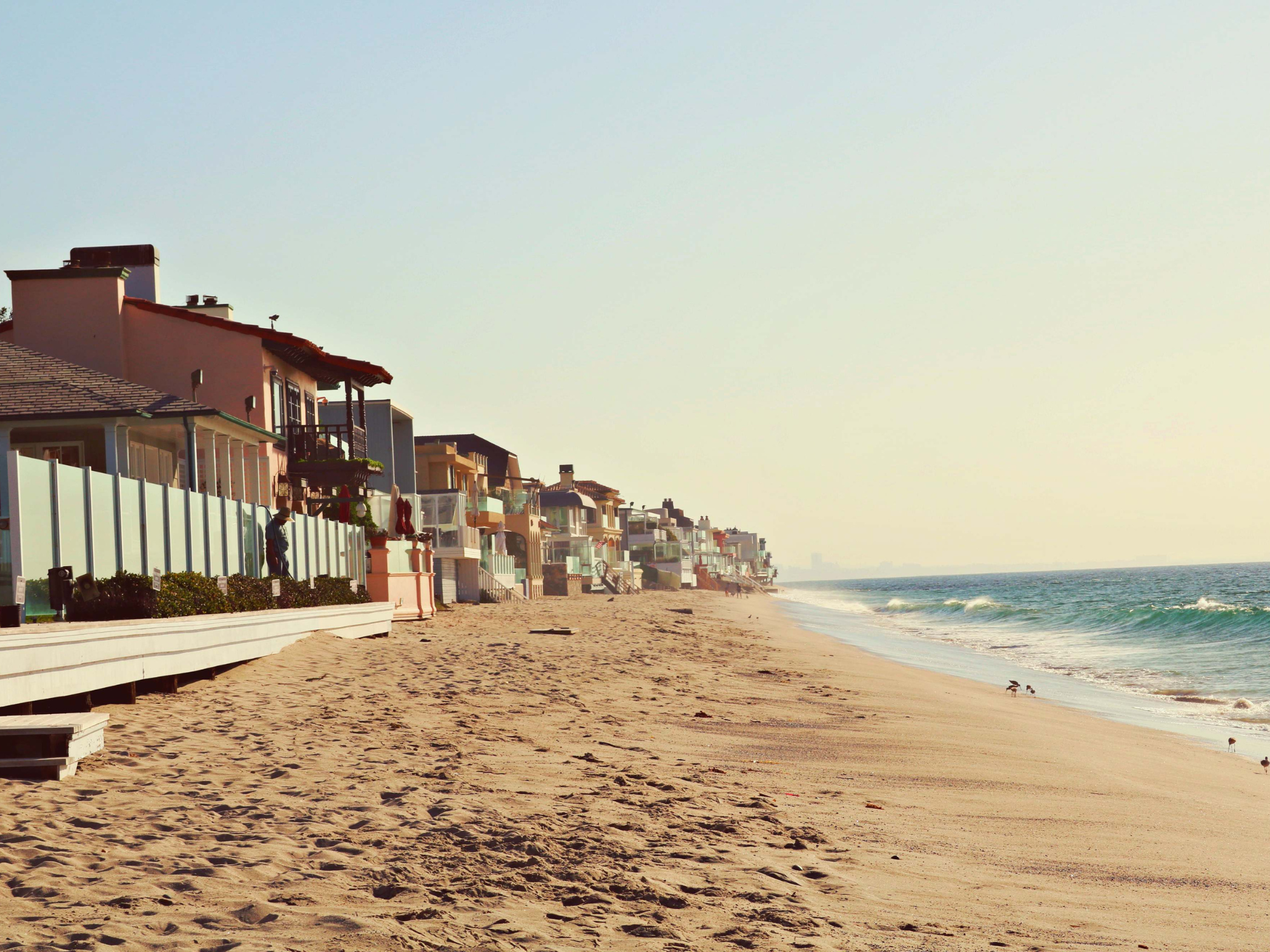 Sunlit beach with row of colorful homes along the sandy shoreline and ocean waves rolling in under a clear sky.