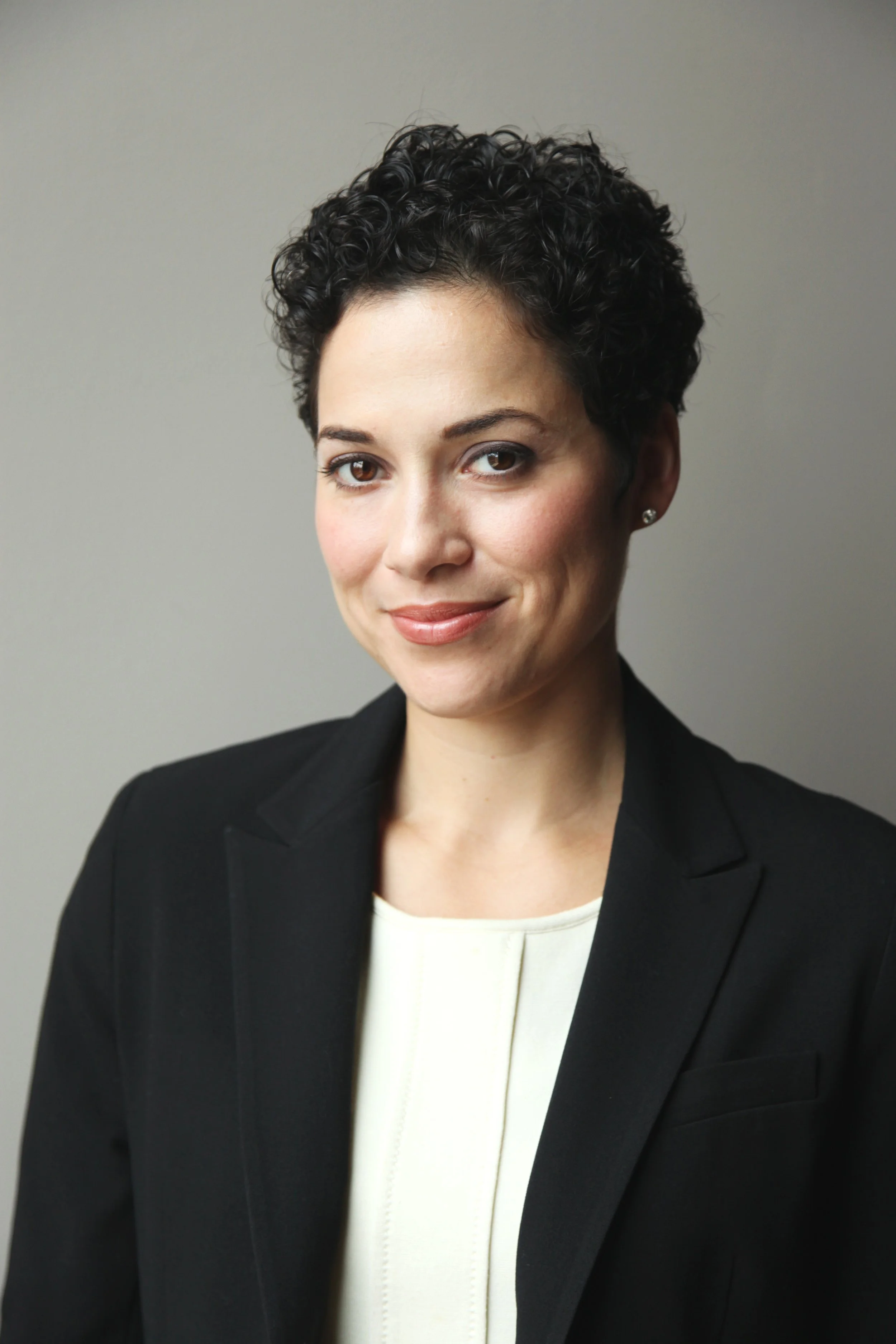 Portrait of a woman with short curly dark hair wearing a black blazer and a cream-colored top, smiling softly against a plain gray background.