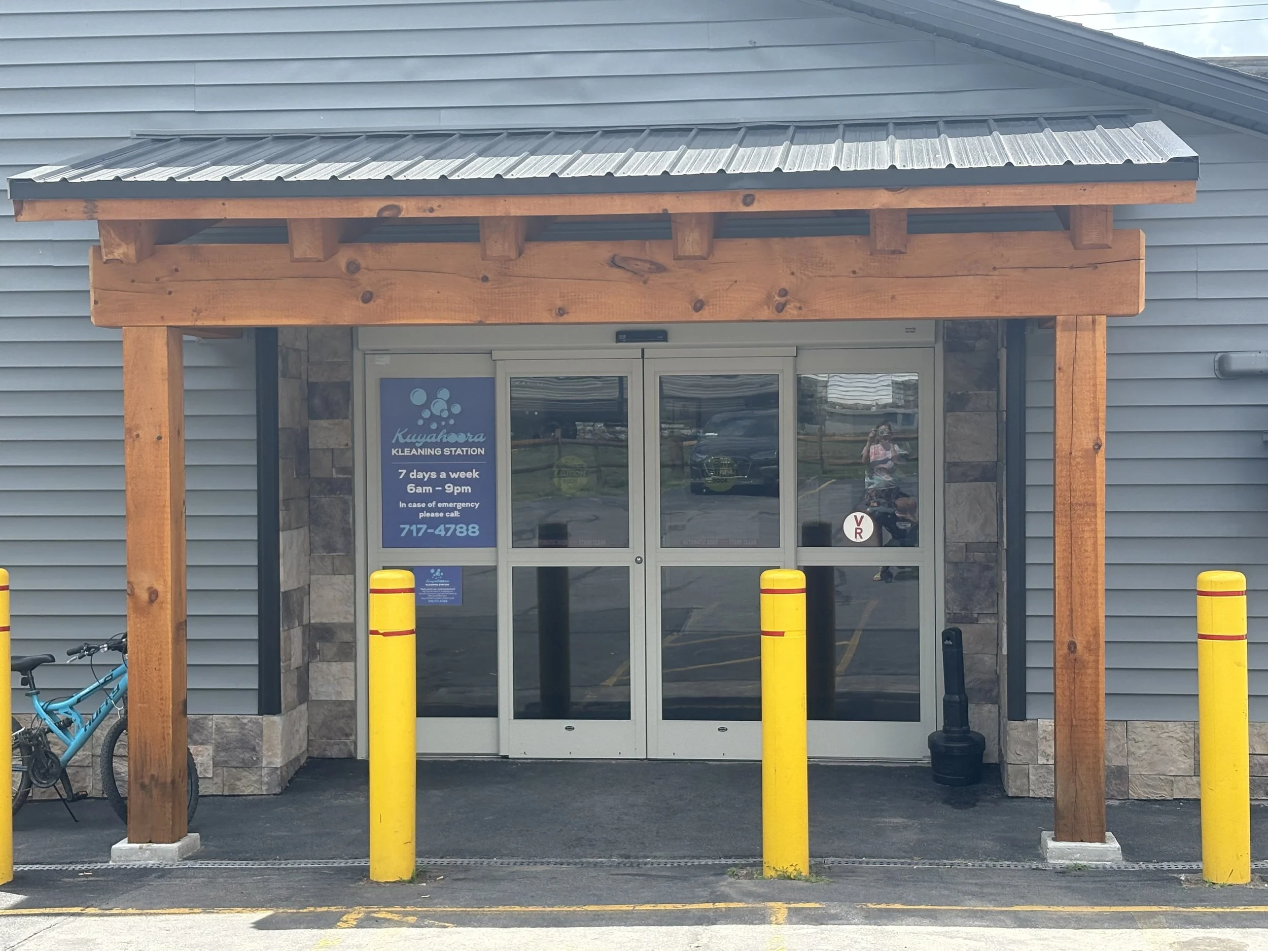 Outside of a laundromat entrance with a wooden canopy, glass doors, yellow parking bollards, and a bicycle on the left.