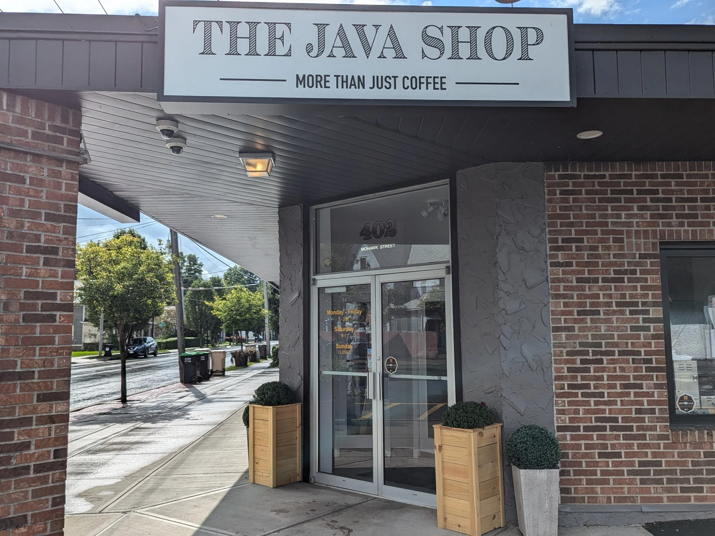 Exterior view of The Java Shop storefront in Herkimer NY with sign reading 'More Than Just Coffee', brick walls, potted plants at the entrance, and a sidewalk with trees and parked cars along the street.