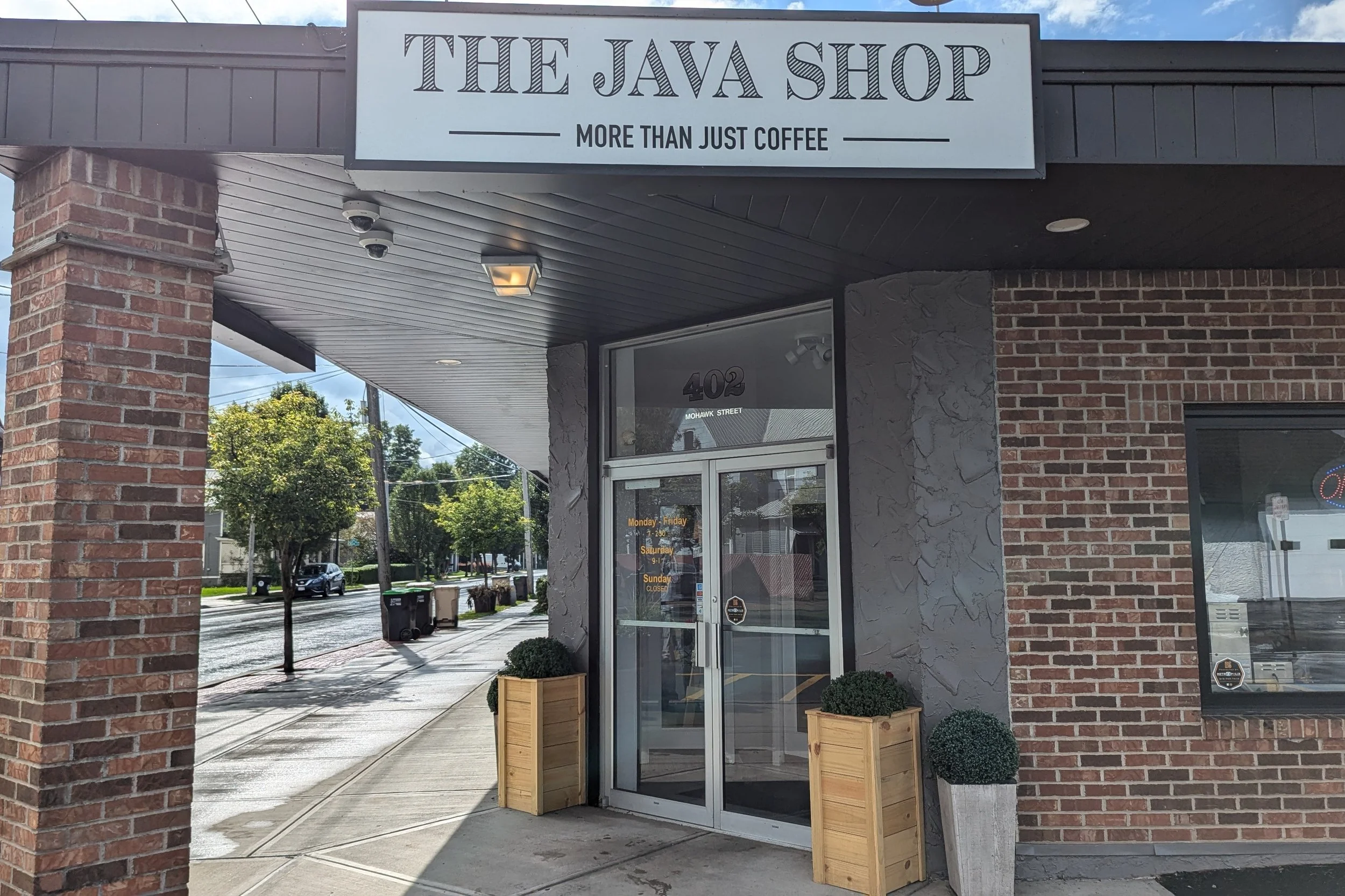The storefront of The Java Shop in Herkimer NY, featuring a sign that says "More Than Just Coffee," with an entrance door flanked by potted plants, and a street scene with trees and parked cars in the background.