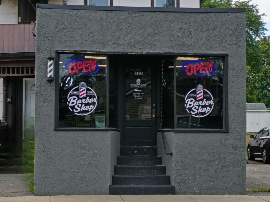 The storefront of a small barber shop with a gray exterior. There are two neon signs that say 'OPEN' in pink and blue, and two circular window decals with the text 'Custom Cutz Barber Shop' and a barber pole graphic. The shop has a black door and ste