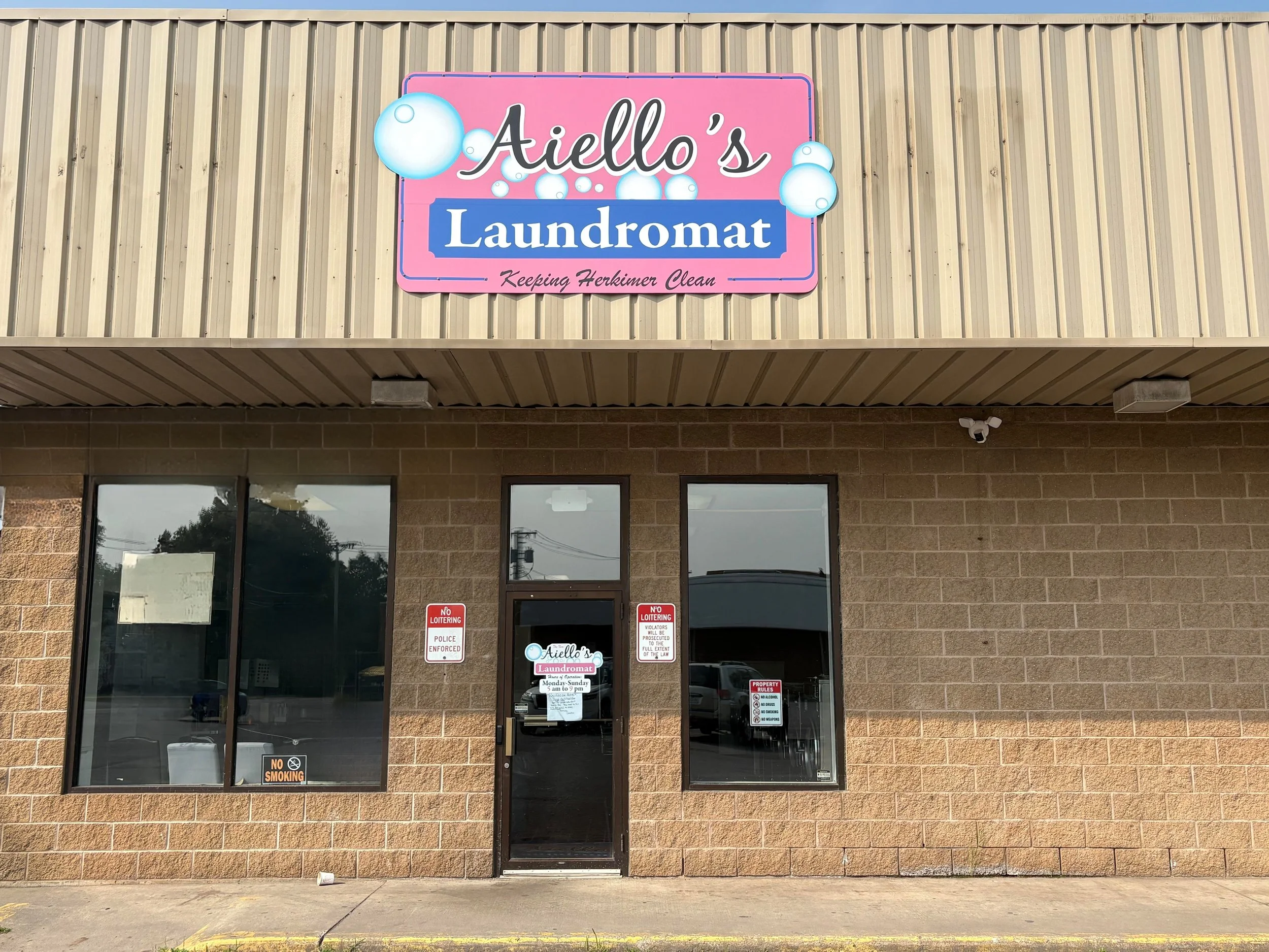 Exterior view of Aiello's Laundromat with a pink and blue sign on a beige brick building. The sign reads 'Aiello's Laundromat' with bubbles design and the tagline 'Keeping Herkimer Clean.' The storefront has three large windows and a glass door with