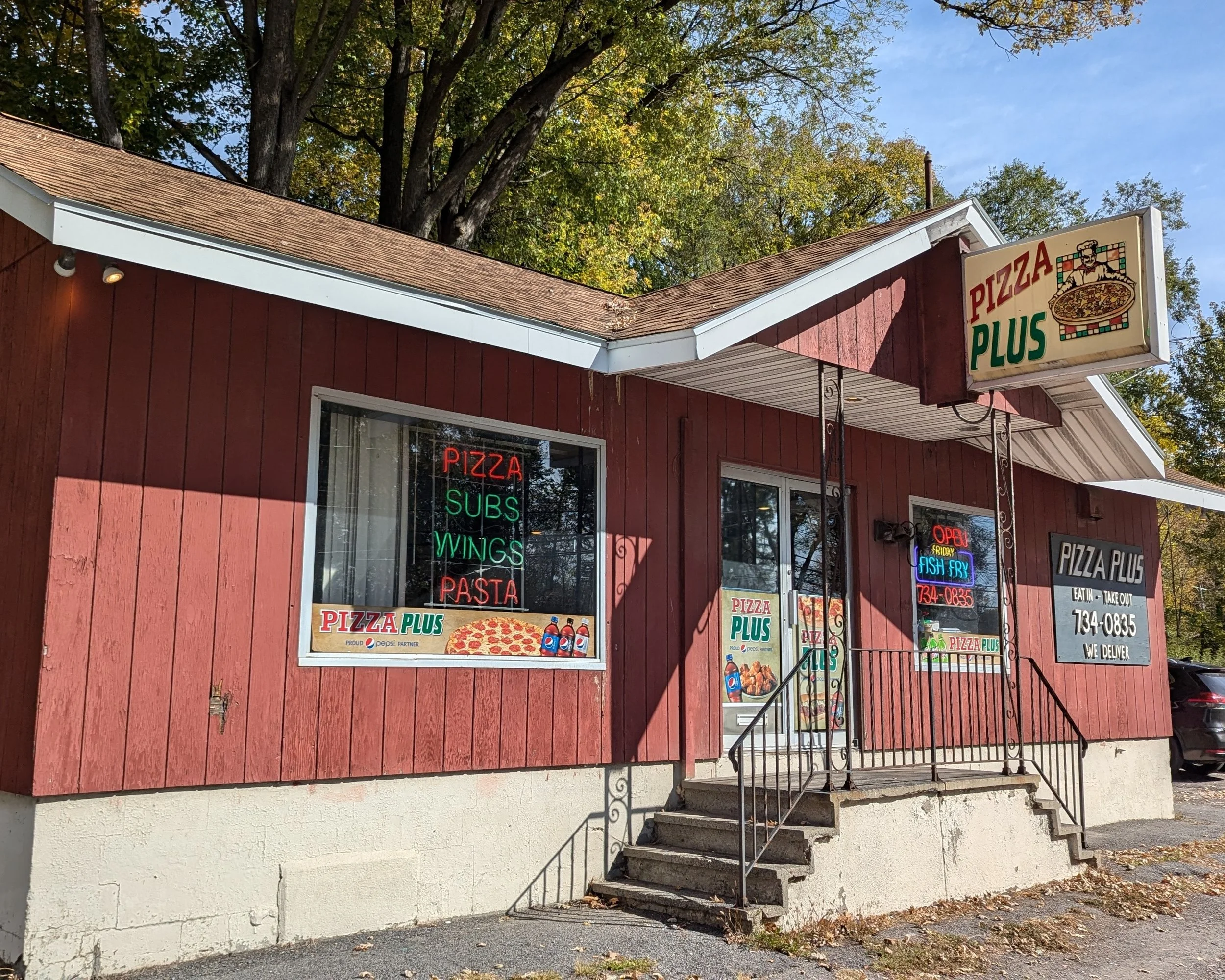 Red small restaurant named Pizza Plus in Utica NY with neon signs listing pizza, subs, wings, and pasta, and a larger sign above the entrance. The building has a steps leading up to the door, surrounded by trees with autumn leaves.