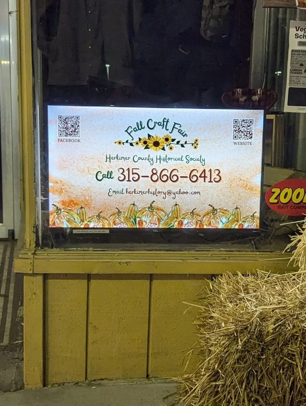 ProBoard digital screen in a storefront window showing a local event promotion ad, beside straw bales and a yellow counter