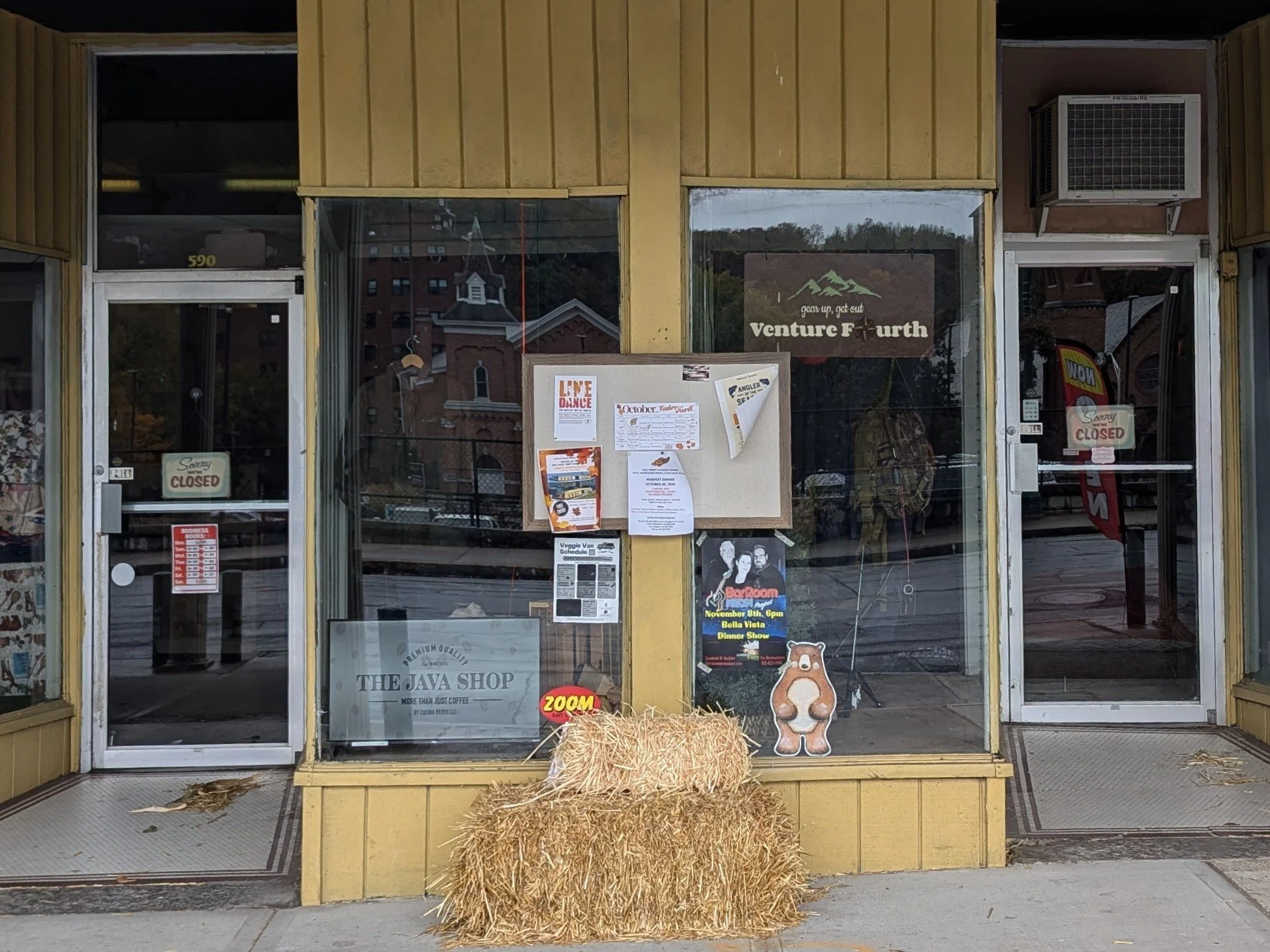 The storefront of The Java Shop in Herkimer NY with yellow exterior, glass windows, and doors. There is a bale of hay outside with a cartoon bear cutout and various posters and signs on the window.
