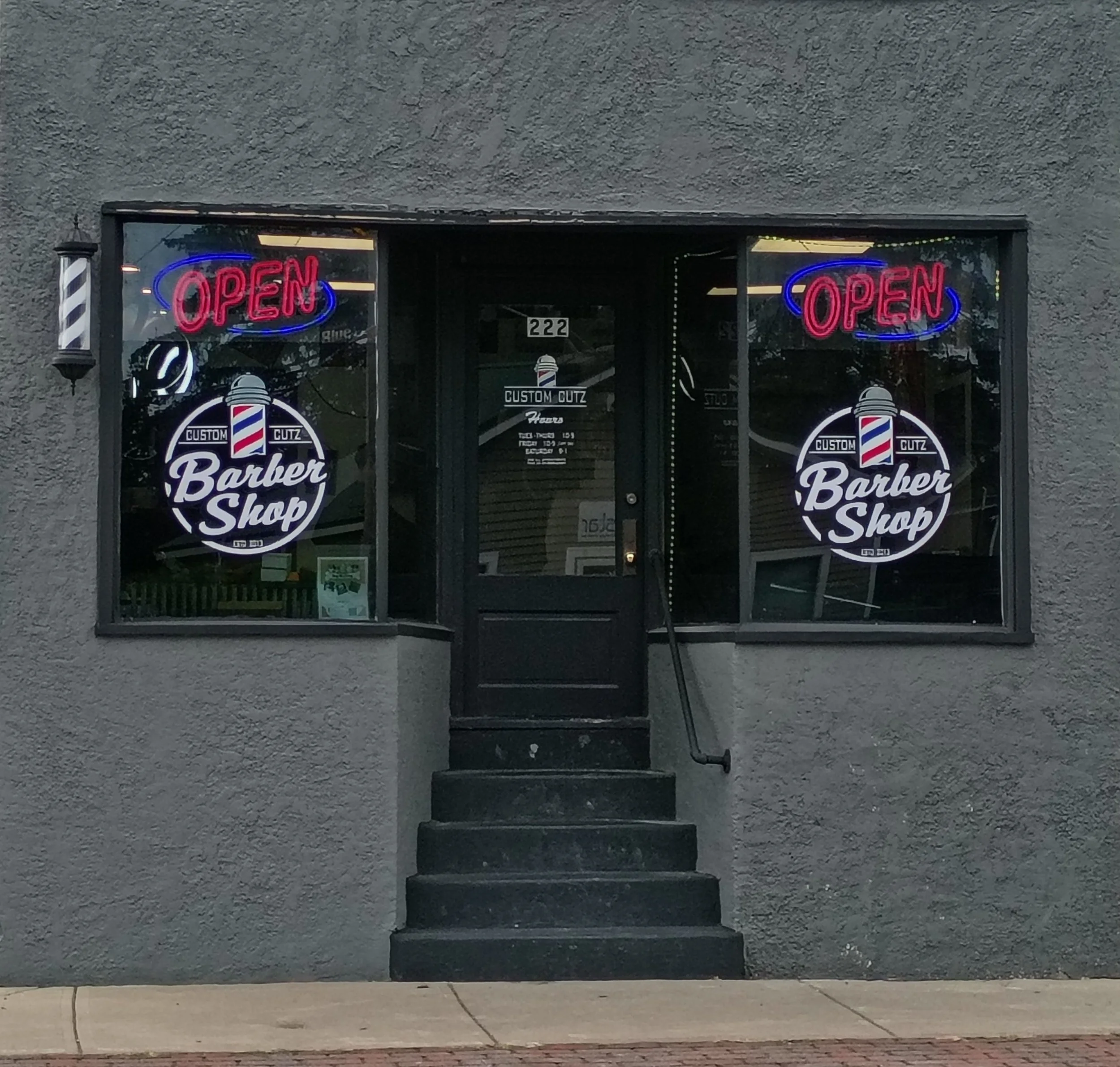 Exterior of a barber shop with gray textured walls, black door with the number 222, two large glass windows displaying neon 'OPEN' signs and barber shop logos, a small black staircase and a black handrail leading to the door, with a sidewalk in front.