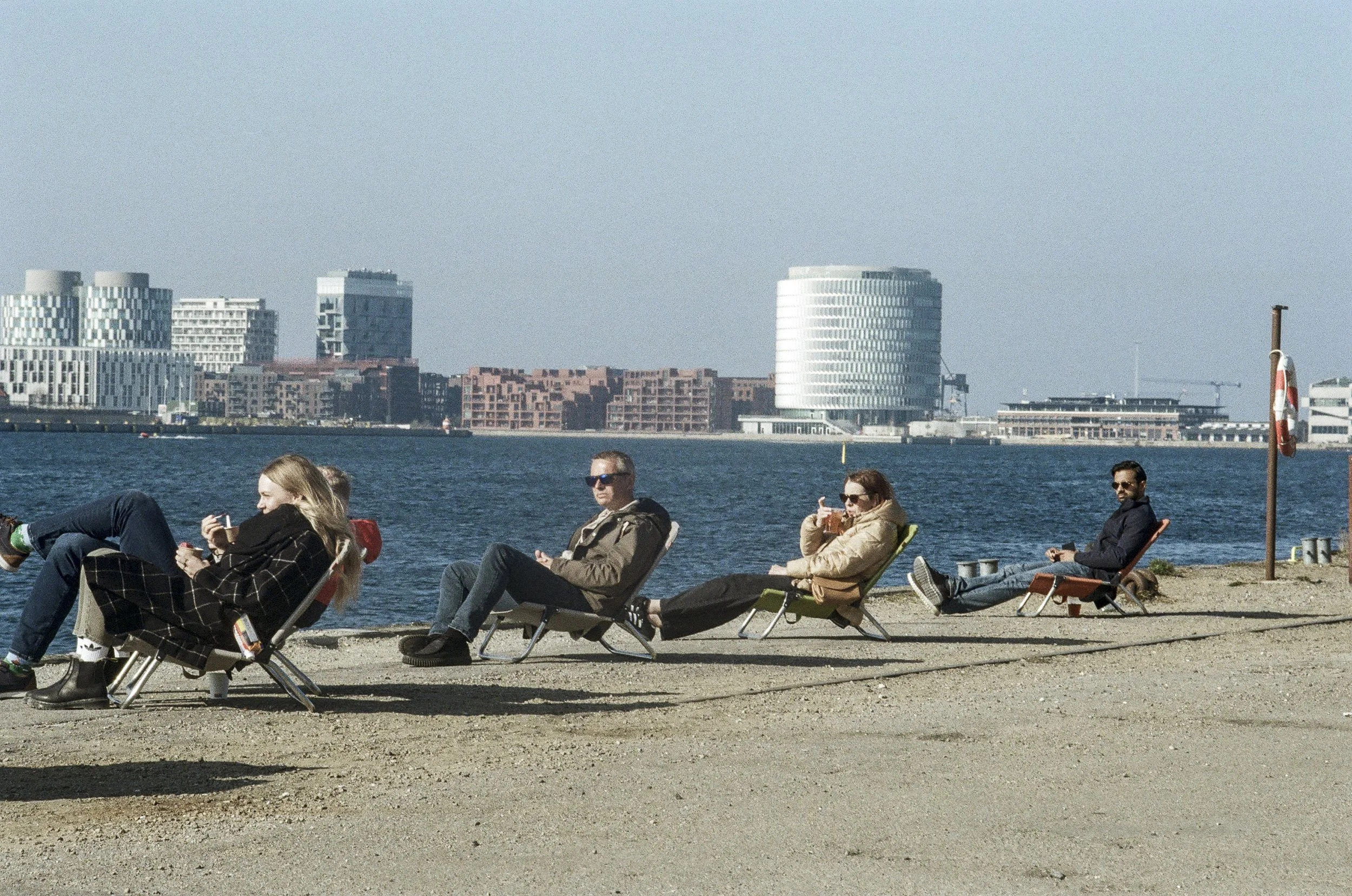 People relaxing on lounge chairs by a water body with modern buildings in the background.