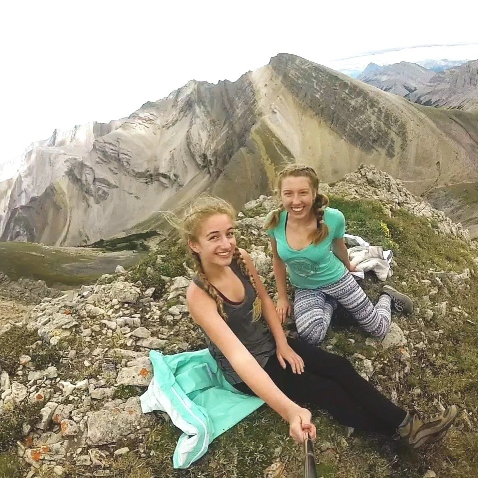 Two young women with braids sitting on rocky mountain terrain, smiling at the camera, with scenic mountains in the background.