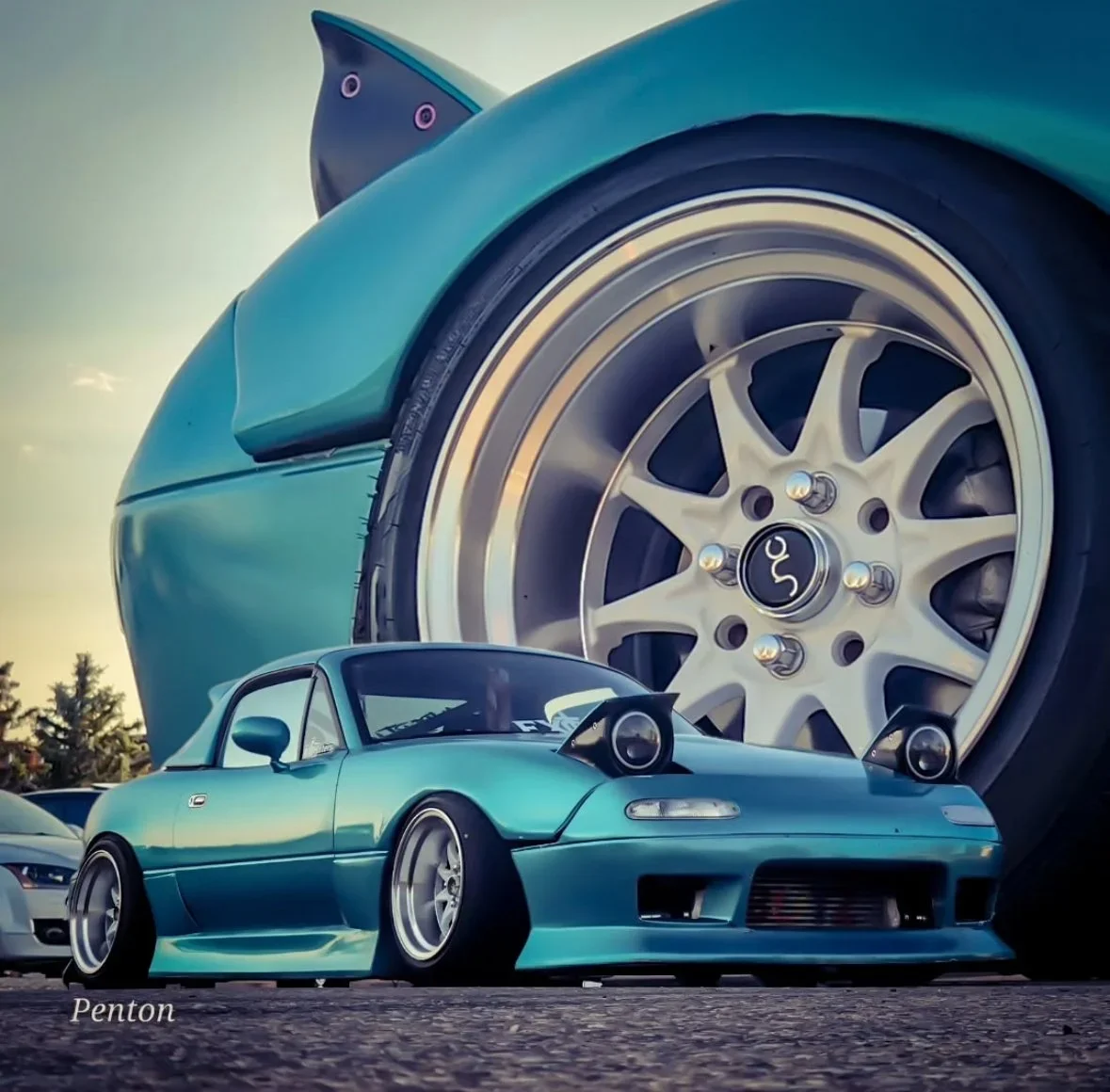 A close-up of a vintage light blue sports car with a lowered stance, wide tires, and custom wheels, parked on a street with a large anime-style illustration of a teal car's wheel and body in the background.