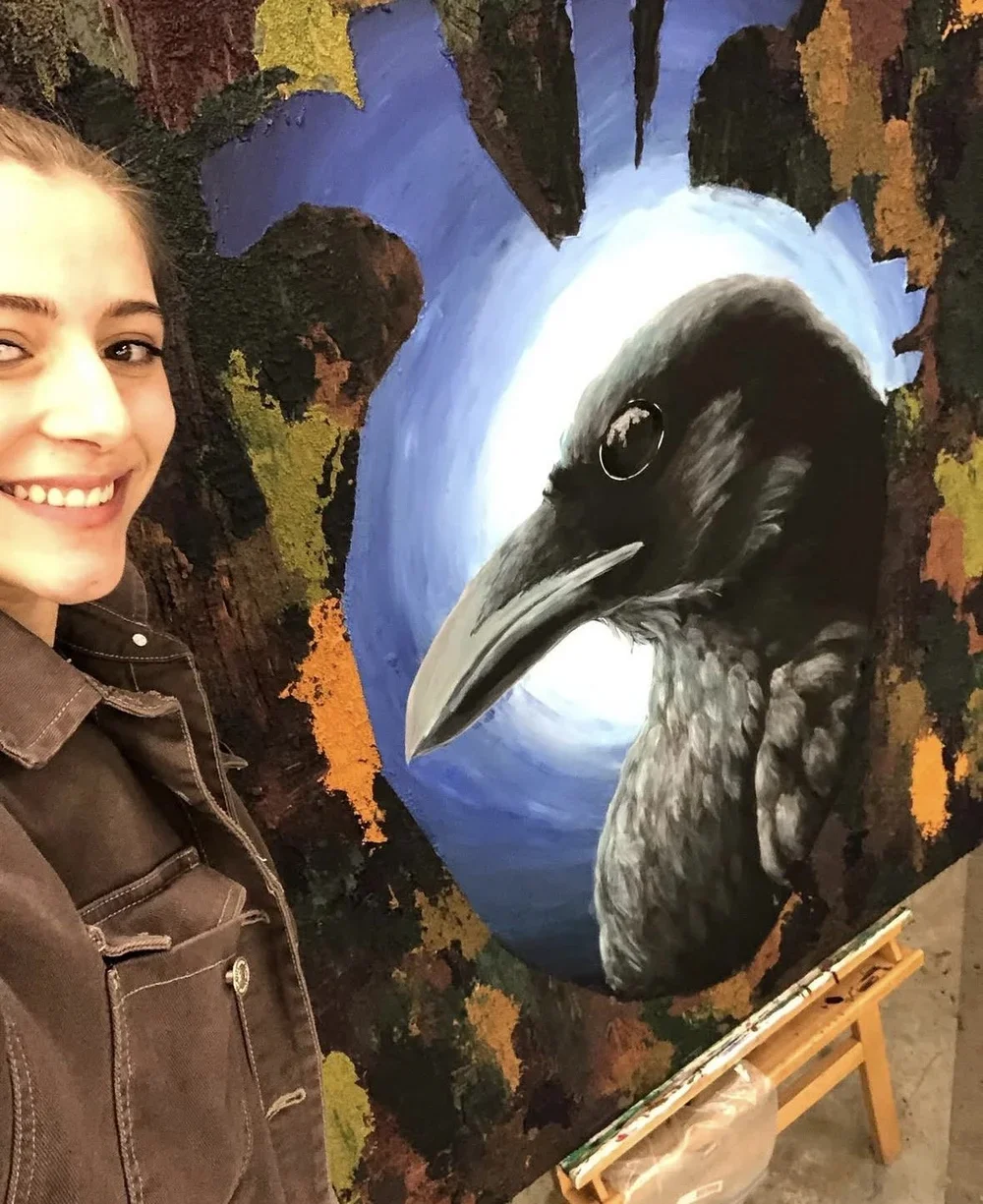 A woman smiling standing next to a colorful painting of a raven with autumn leaves in the background.