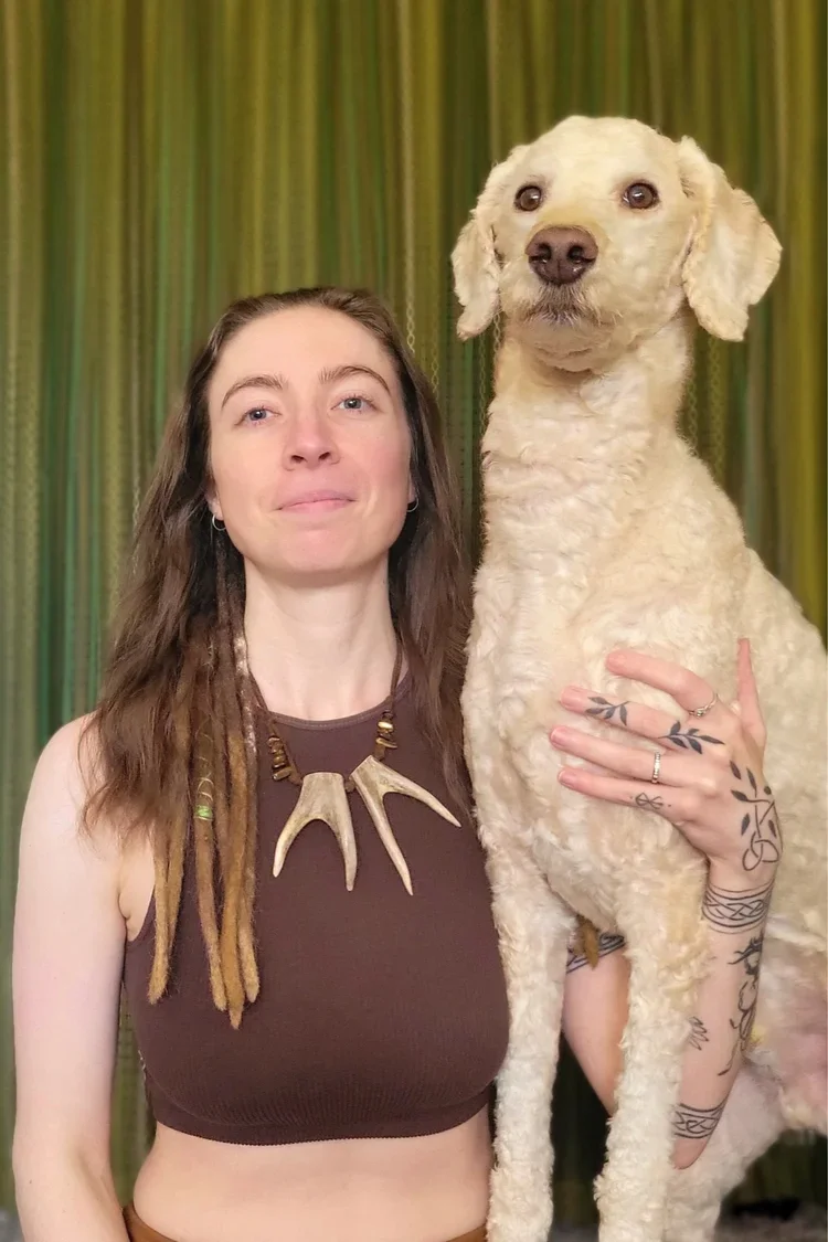 A woman with long, wavy hair and tattoos on her hand holds a large, curly-haired dog with tan fur. The woman is wearing a brown sleeveless top and a necklace made of antlers. The background features green curtains.
