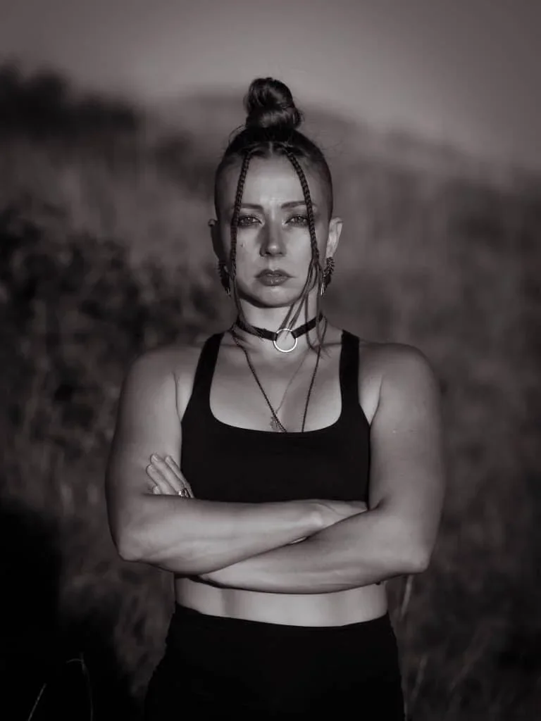 Black and white photo of a young woman with braids and a top knot hairstyle, standing outdoors with her arms crossed, wearing a sleeveless crop top and jewelry, with a blurred natural background.
