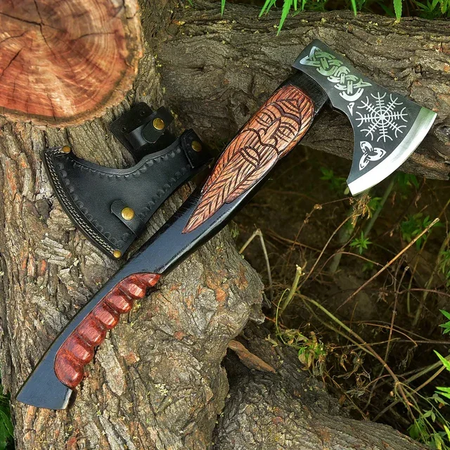 A decorative axe with a carved wooden handle, resting on a tree trunk outdoors. The axe head has Nordic-style engraved designs with snowflakes.