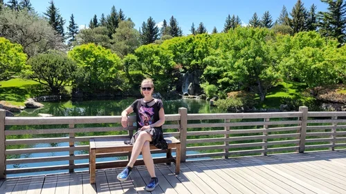 A woman sitting on a park bench by a pond with lush green trees and a small waterfall in the background.