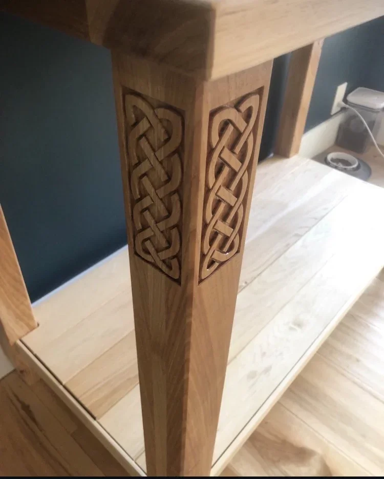 A wooden table leg with intricate carved knot patterns in a warm finish, part of a piece of furniture with a light-colored wooden surface.