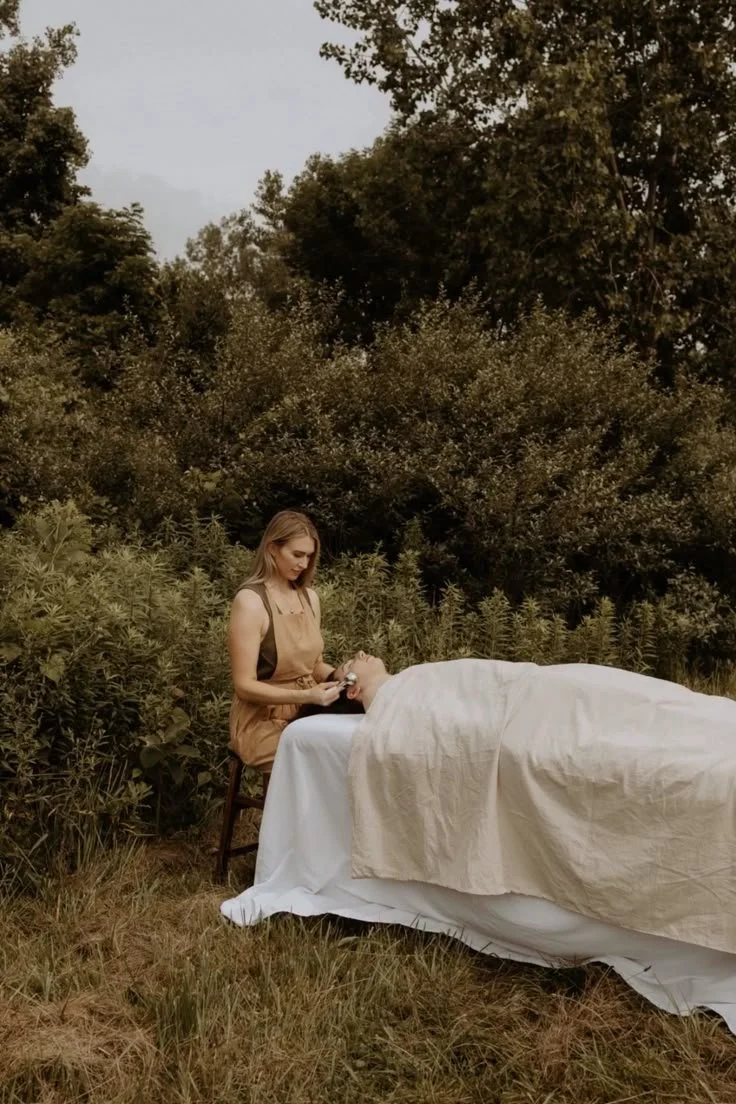 A woman receiving a massage outdoors in a natural setting with trees and grass, lying on a bed covered with white sheets, while a woman in an apron stands beside her.